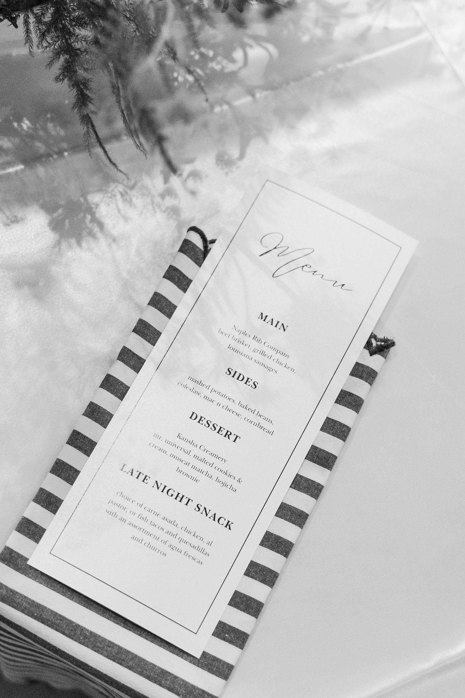 Black and white photo of an elegant wedding menu card on striped napkin with sections for mains, sides, dessert, and late night snack.