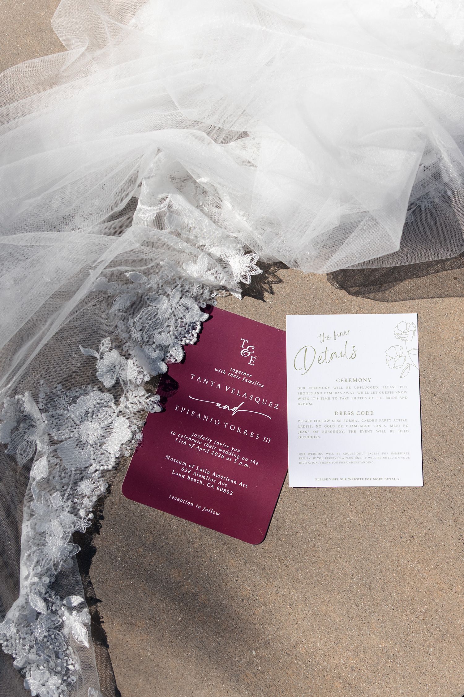Elegant wedding invitation suite in burgundy and white beside a lace bridal gown and veil on a neutral surface.