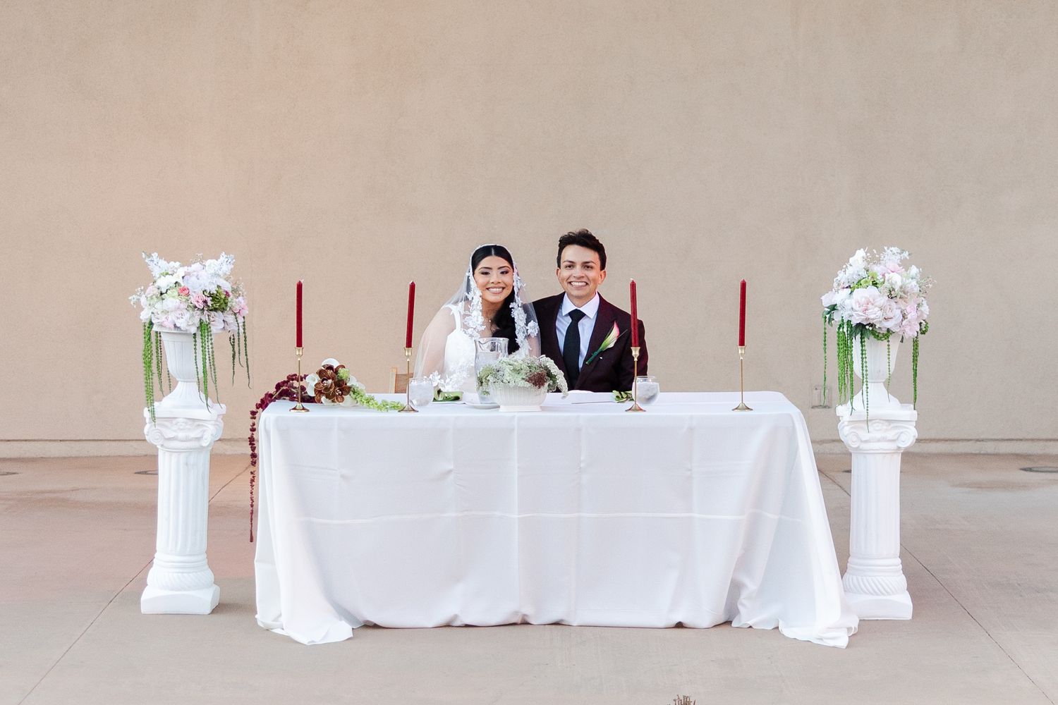 Bride and groom smiling at elegant white sweetheart table with floral arrangements and red candles at wedding reception.