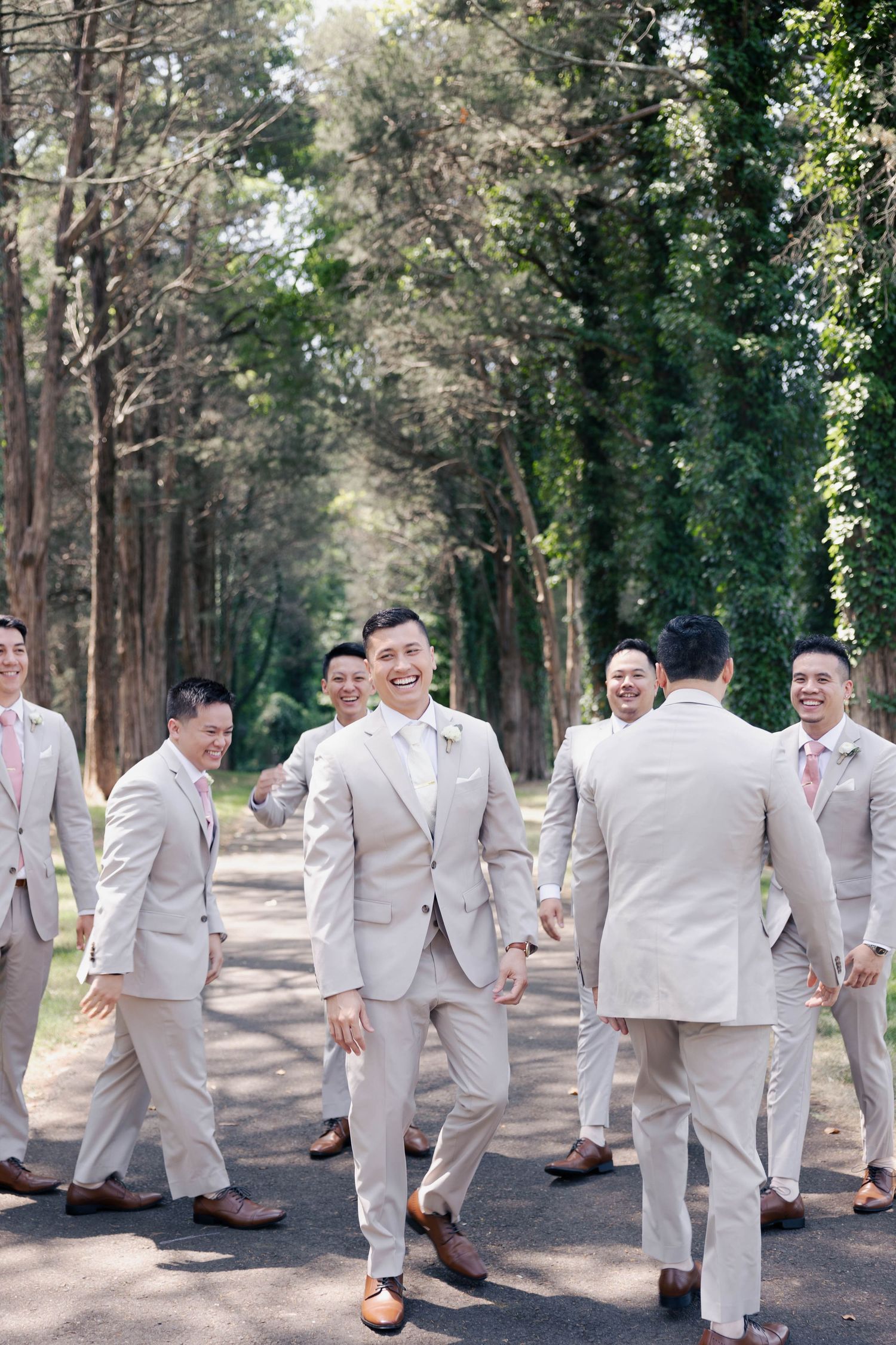 Wedding party members in light suits interact casually on a tree-lined pathway.