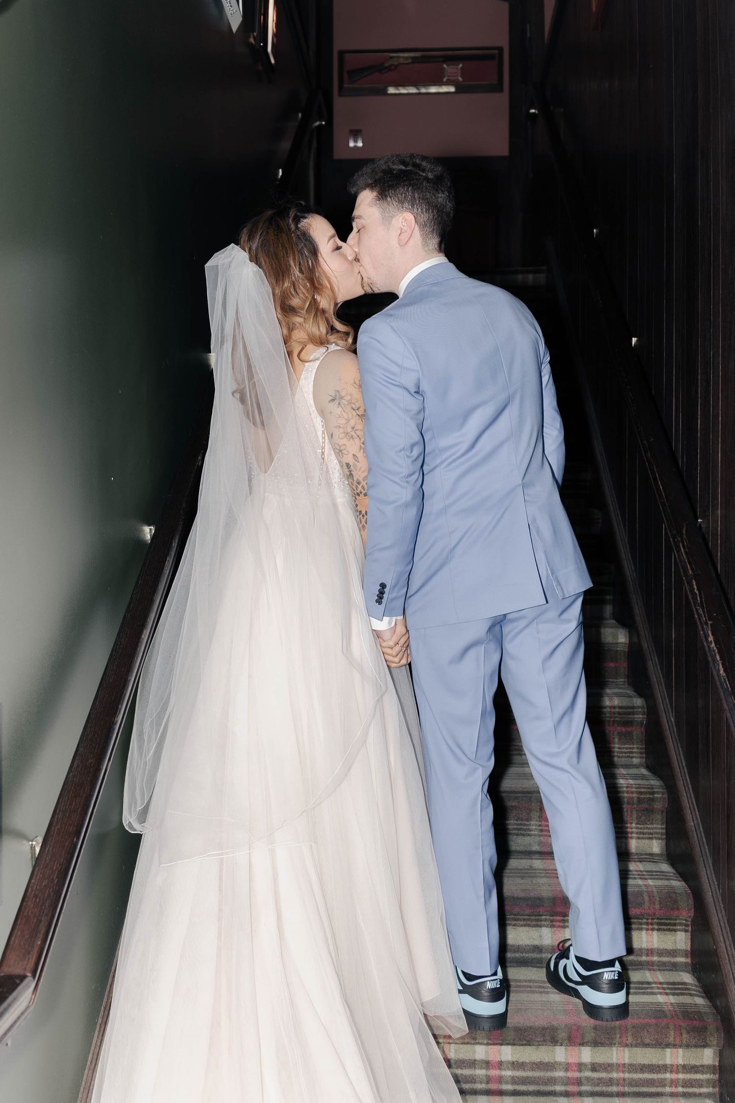 Couple shares a romantic kiss on carpeted stairs, bride wearing flowing white gown and groom in light blue suit.