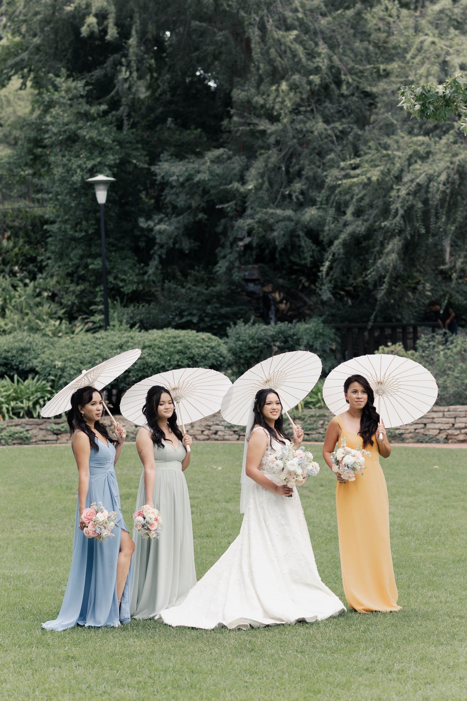 A group in pastel dresses and white parasols poses together on a grassy lawn during an outdoor photo session.