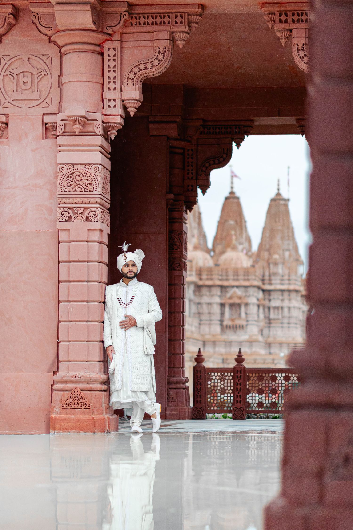 A person in white clothing stands between ornate stone pillars at a traditional Indian temple with carved details.