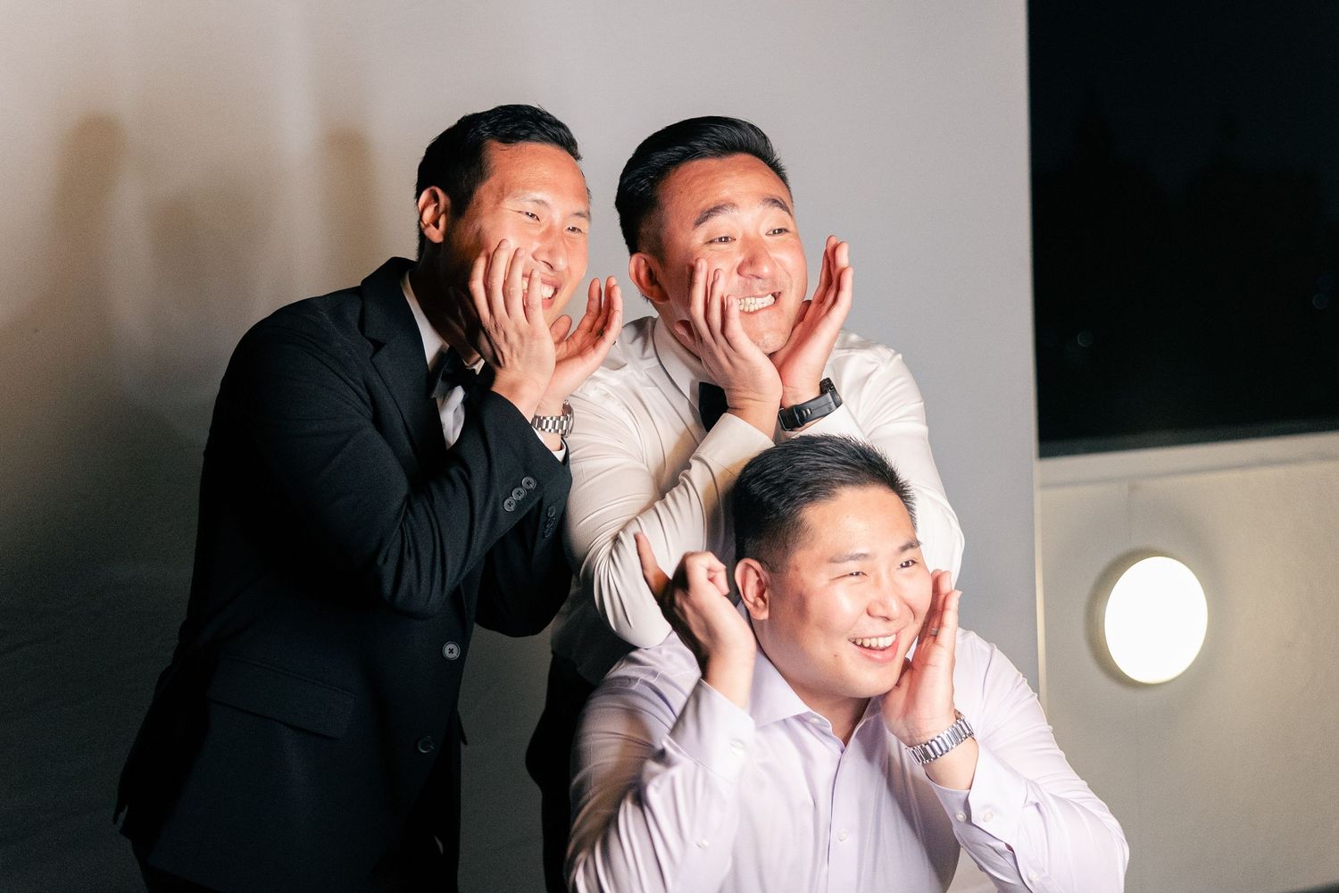 Three men posing playfully at a wedding, making fun expressions with hands on faces, wearing formal attire.