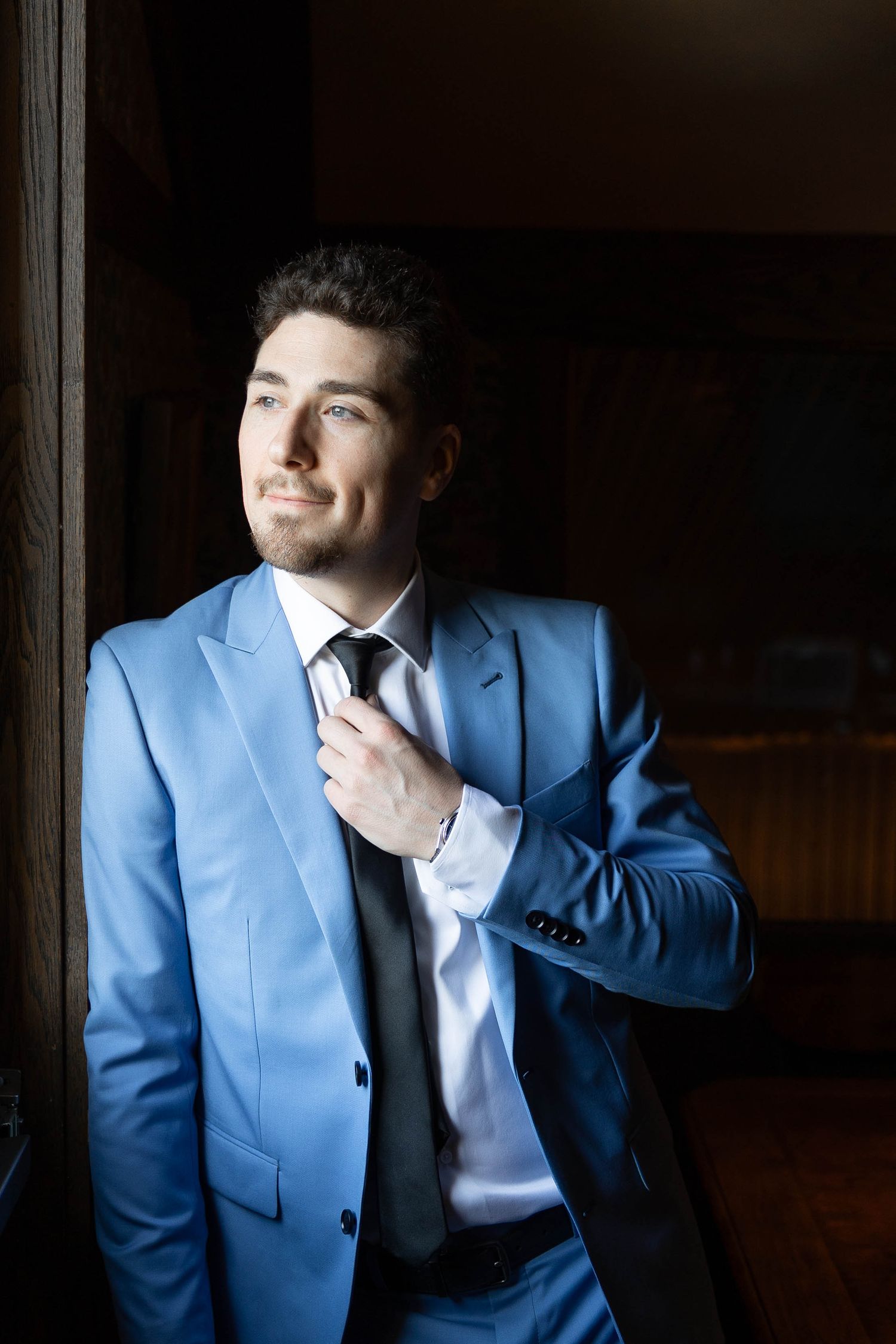 Elegant man in powder blue suit adjusts tie while standing near window with dramatic lighting.