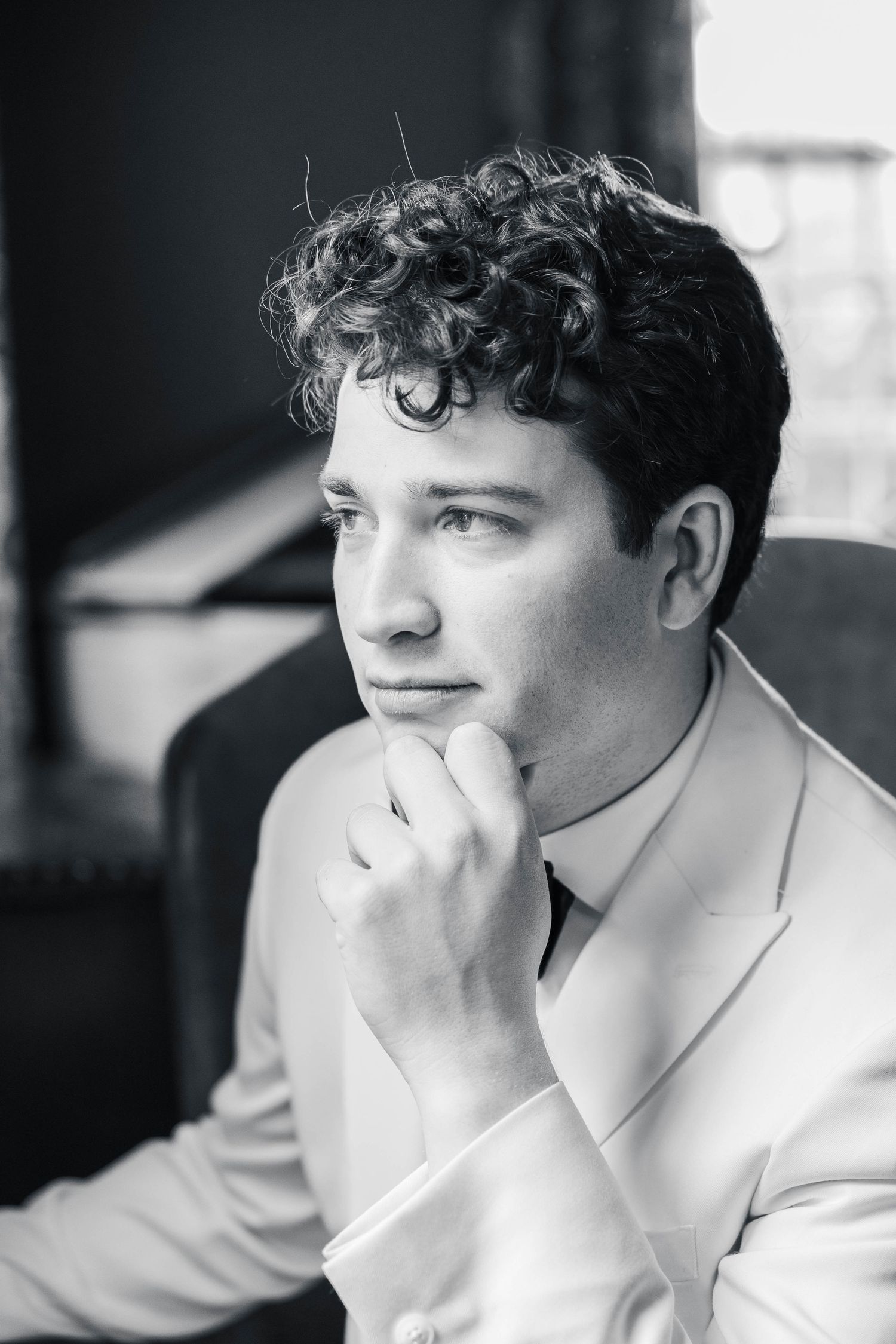 A black and white portrait of someone in a light colored suit jacket with curly dark hair in a contemplative pose.