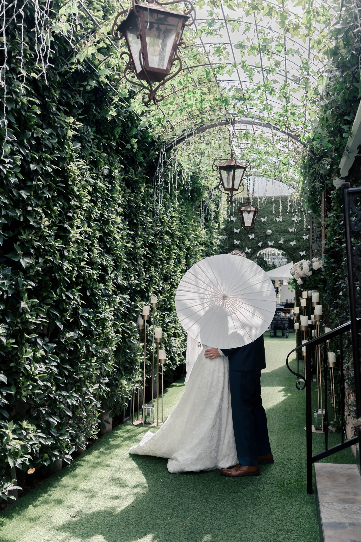 A couple in wedding attire poses with white parasols under a lush green garden archway with vintage lanterns.