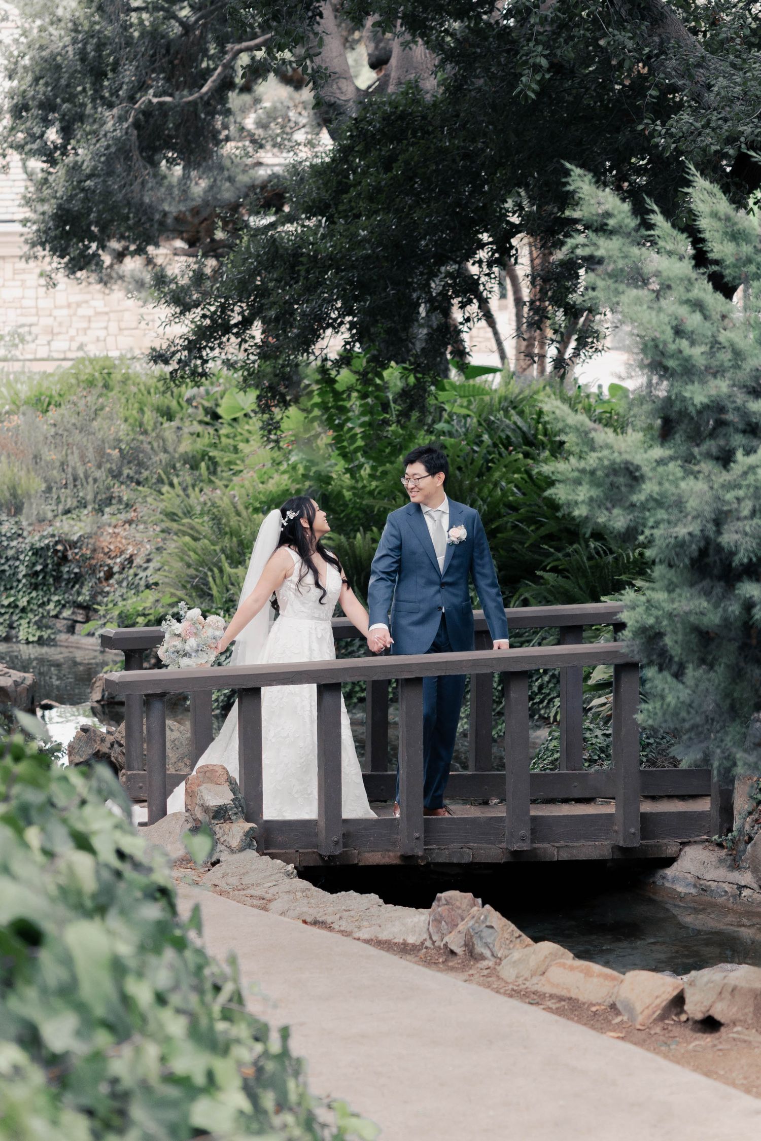 A couple shares romantic moments on a rustic wooden bridge surrounded by lush greenery in a garden setting.