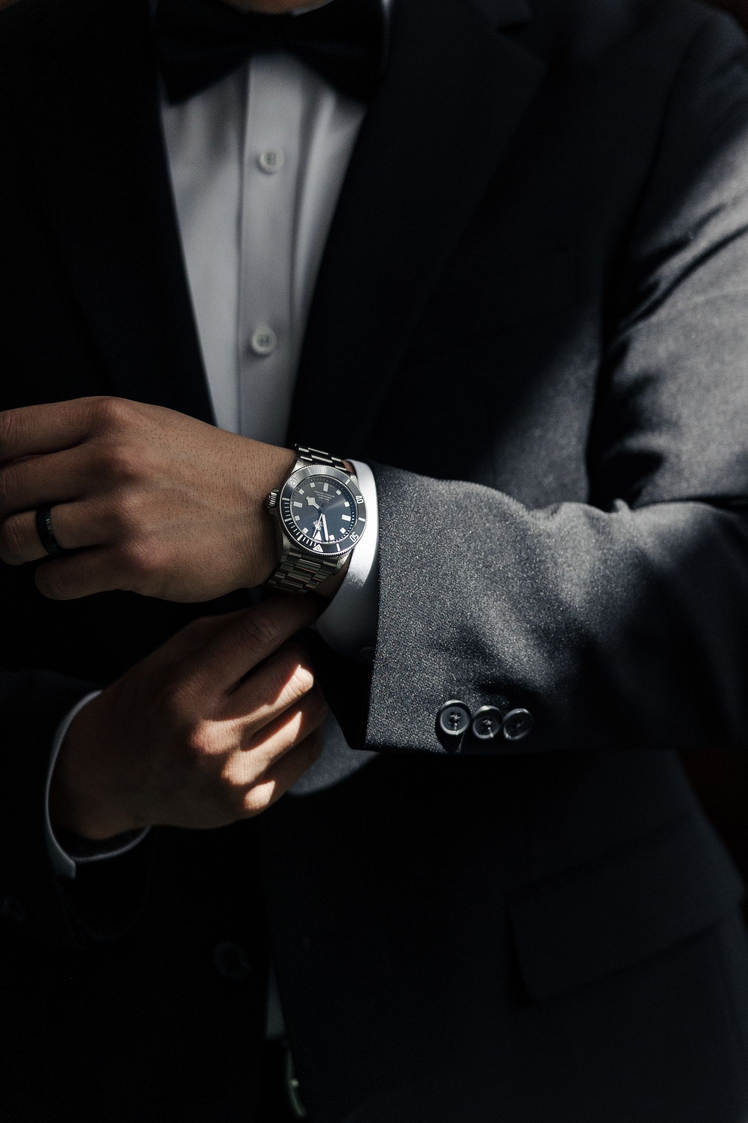 Man in black tuxedo adjusting silver watch on wrist, wearing ring, elegant formal attire with bow tie.