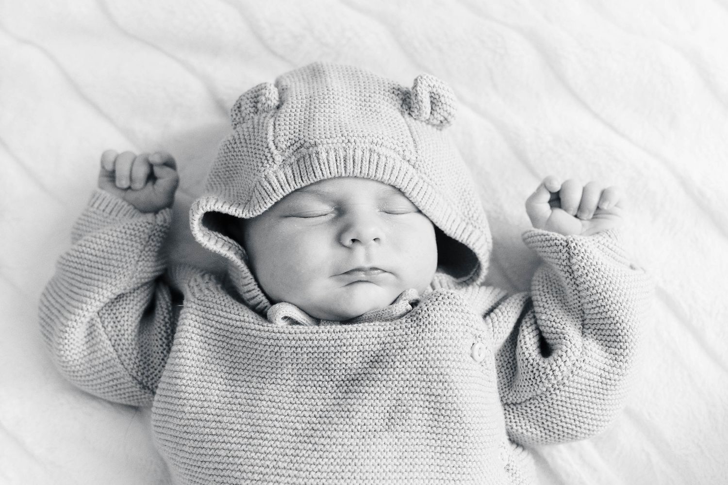 Closeup of a sleeping infant wearing a knit beanie against white textured fabric.