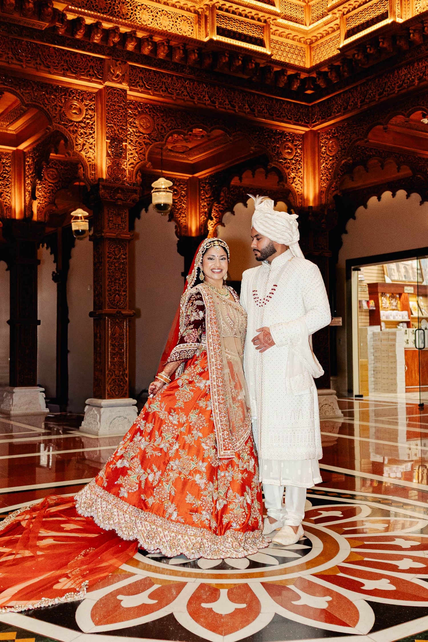 A couple in traditional Indian wedding attire poses in an ornate palace hall with intricate gold details and patterned floor.