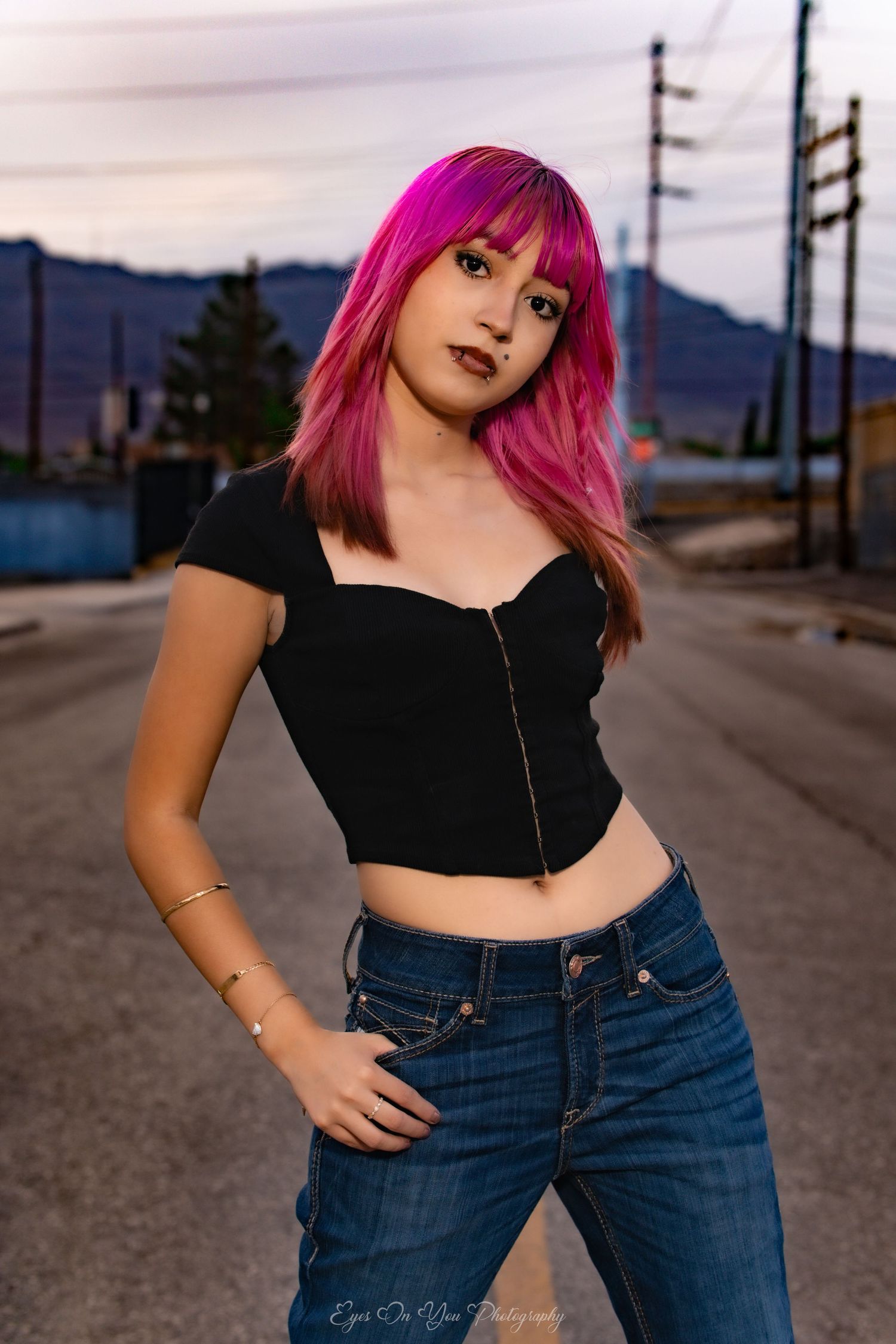 Person with bright pink hair poses in black top and jeans on an empty street at dusk.
