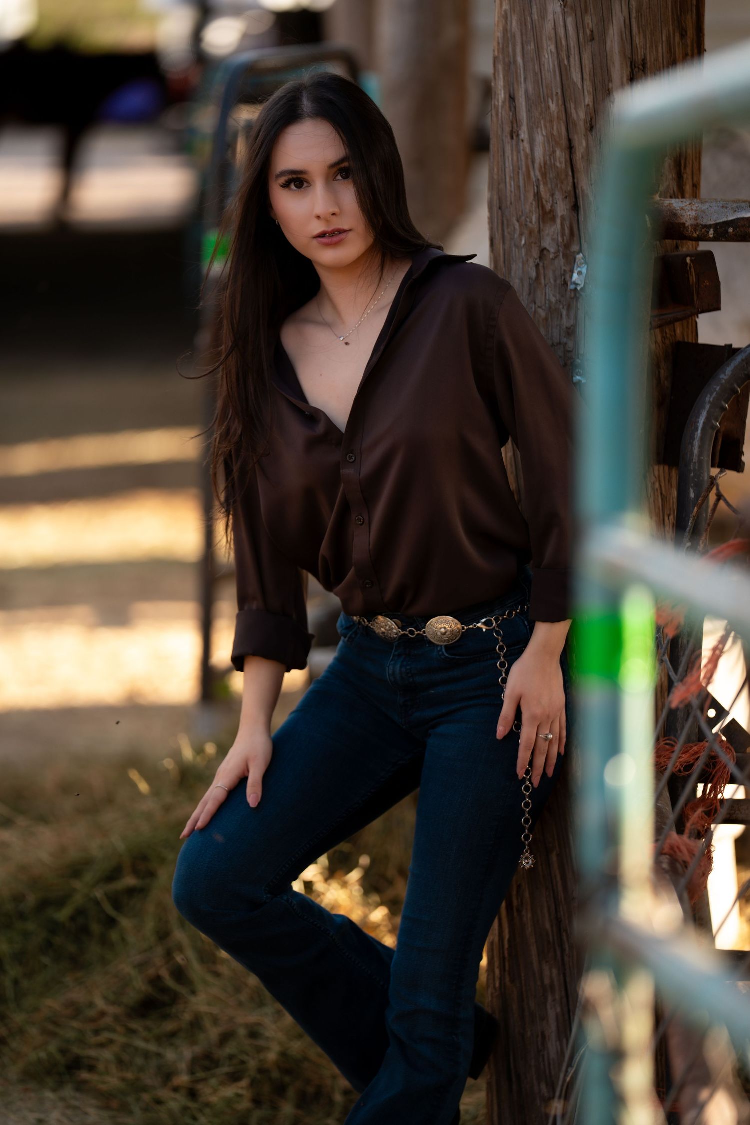A person in a black top and jeans poses casually against a tree trunk in an outdoor setting during golden hour.