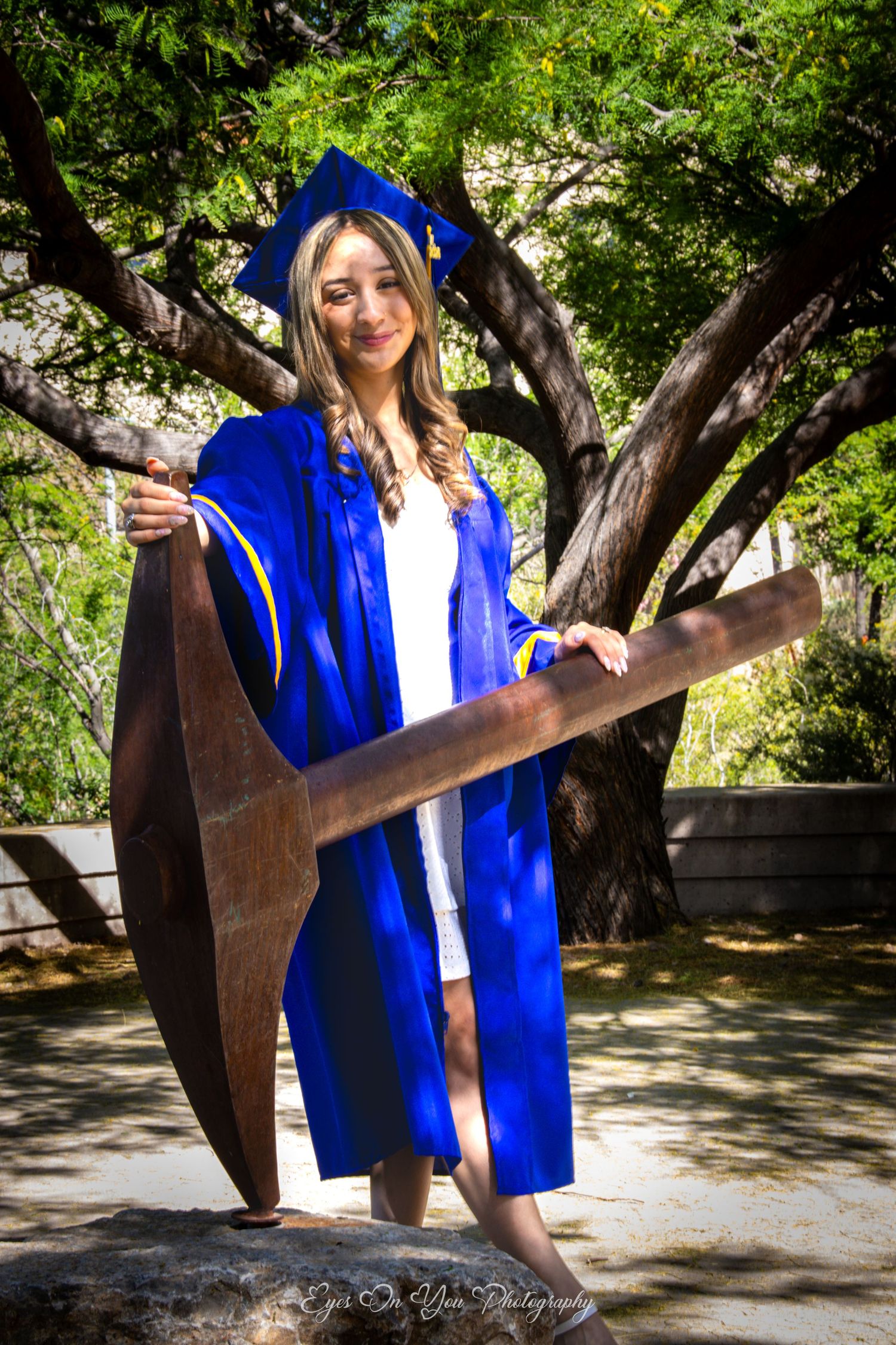 Graduate in blue regalia poses with modern metal sculpture outdoors.