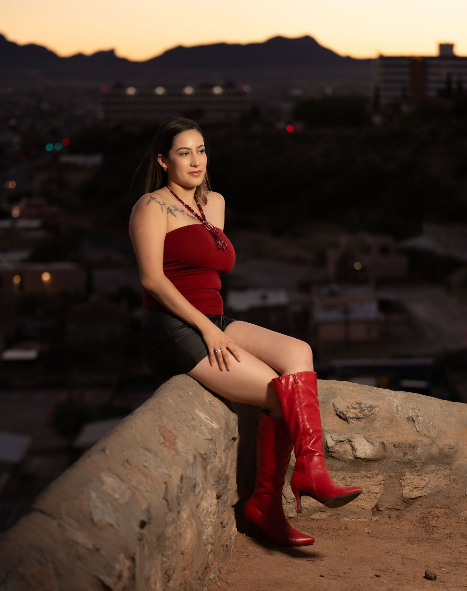 A figure in a red dress and boots poses on a rocky ledge at sunset against an urban backdrop.
