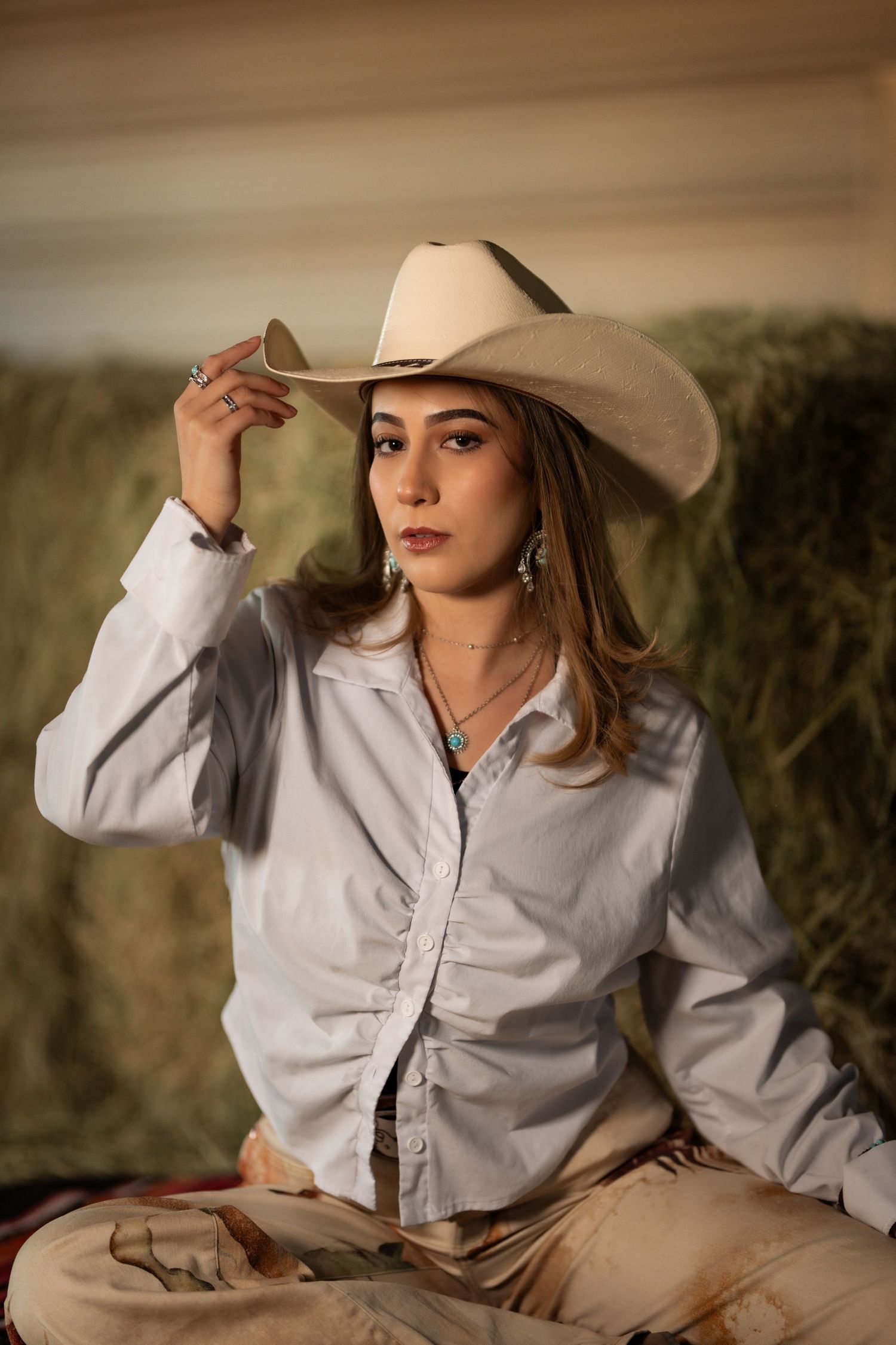 A person in western attire with a white cowboy hat and button-down shirt poses outdoors at golden hour.