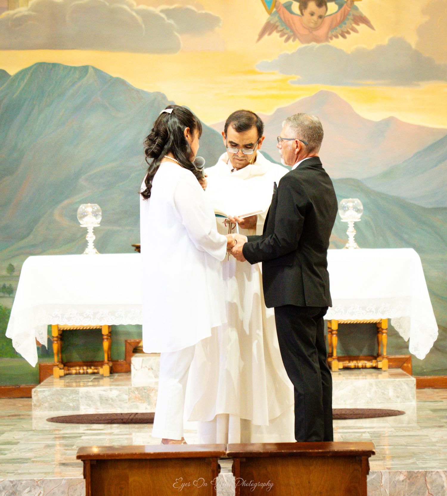 A wedding ceremony takes place on a raised platform with mountains and a golden sunset painted on the backdrop behind.