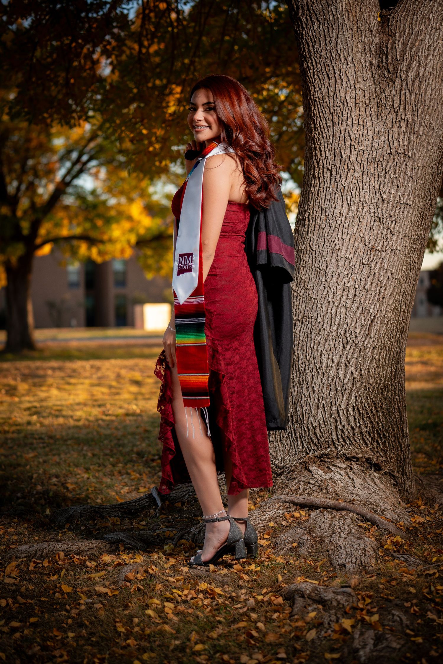 A figure in a burgundy velvet dress and black jacket poses against autumn trees in a park setting at sunset.