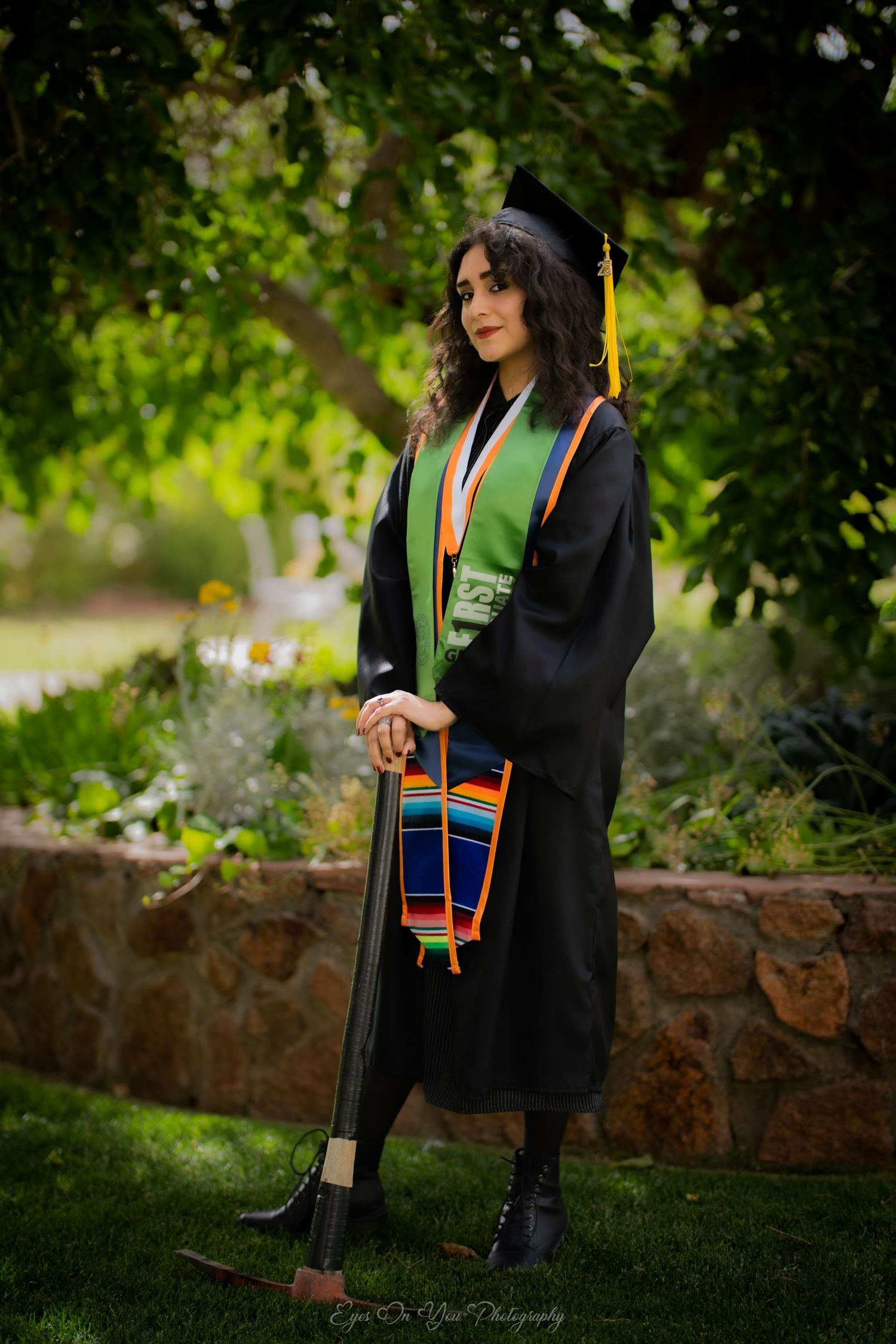 Graduate in cap and gown posing against stone wall with decorative stoles.