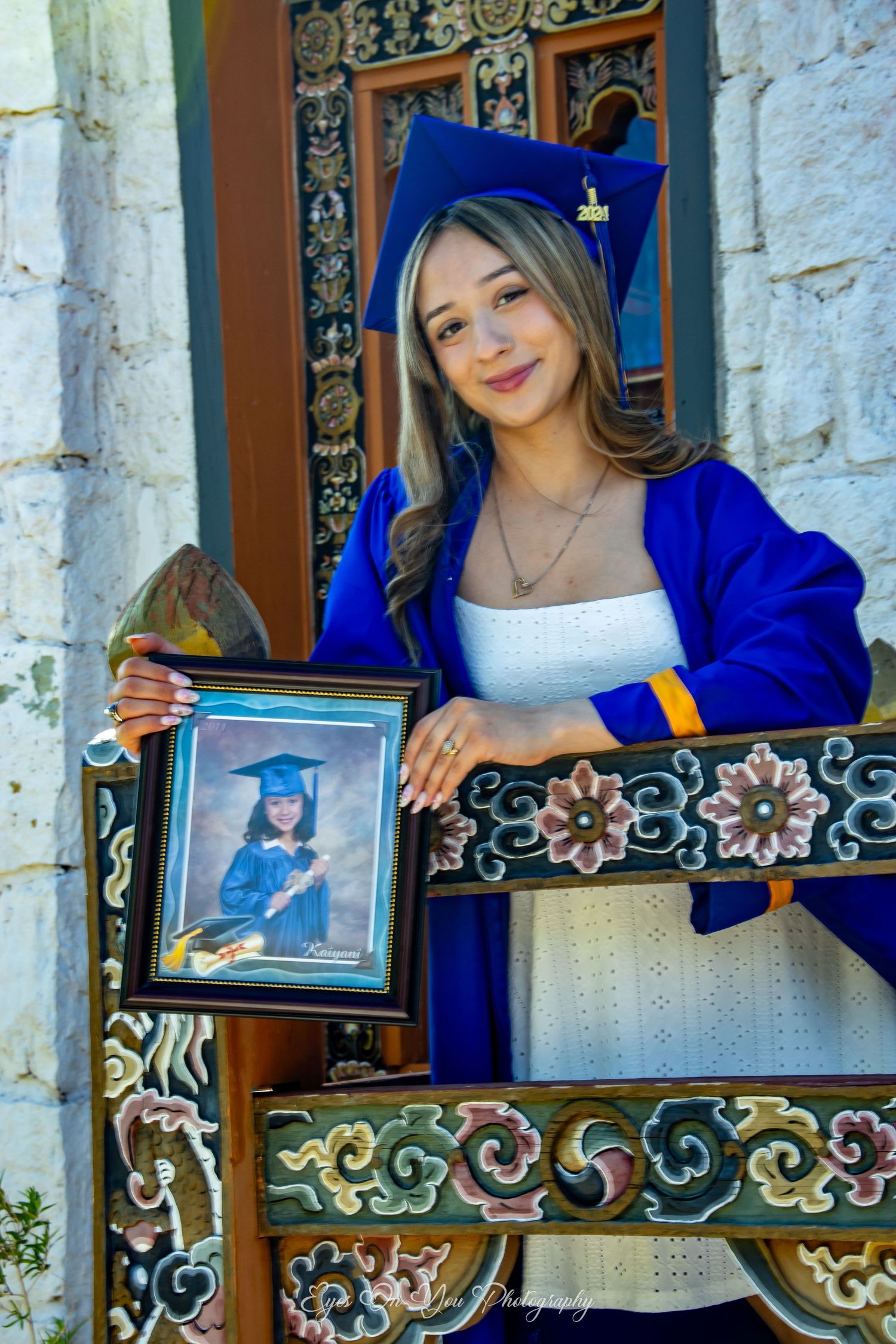 Graduate in blue cap and gown poses with framed photo outdoors.