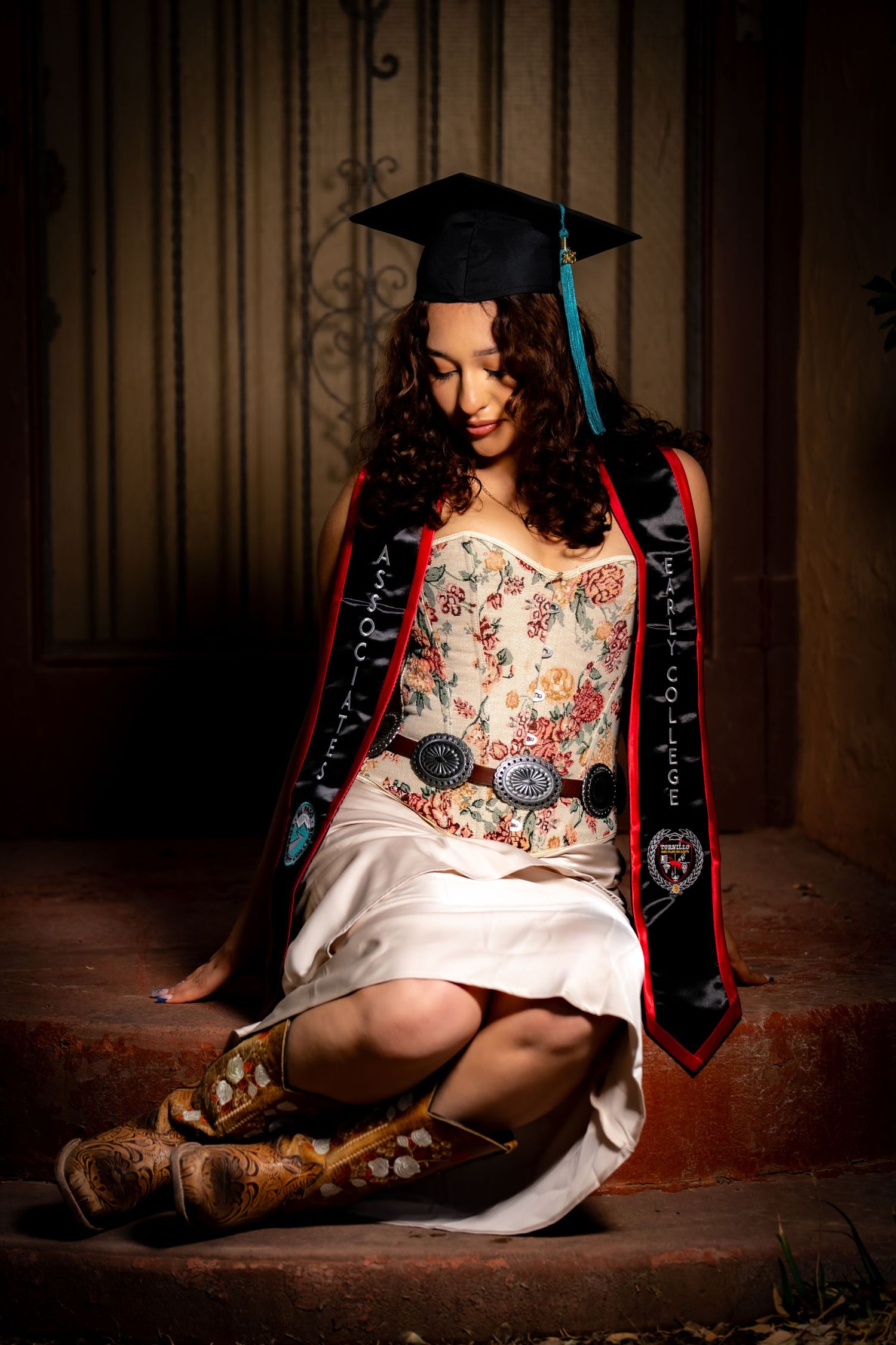 A young woman in graduation cap and sash sits on steps wearing a floral dress and cowboy boots.