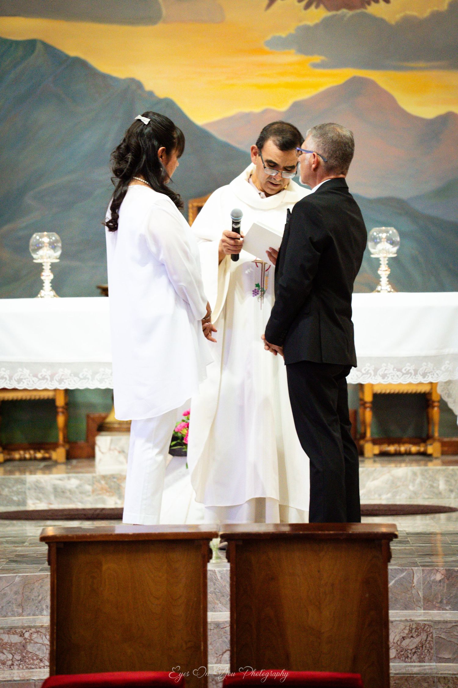 A wedding ceremony takes place with a scenic mountain sunset backdrop as religious officials gather at the altar.