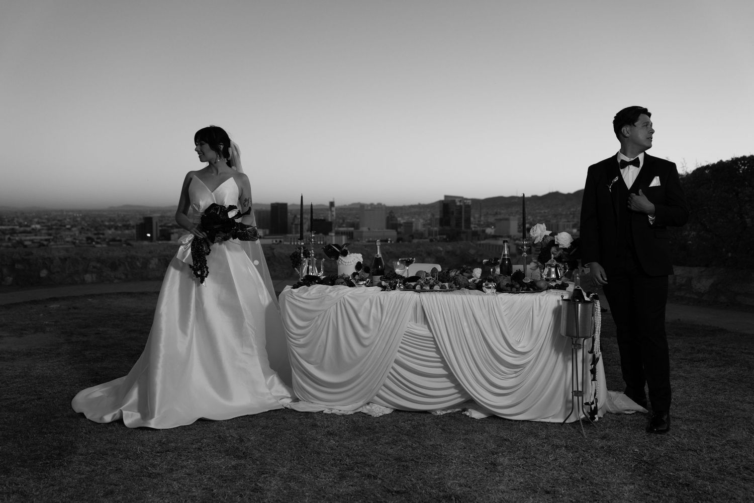 A black and white wedding reception photo showing a couple and an elegantly draped table at an outdoor venue at dusk.