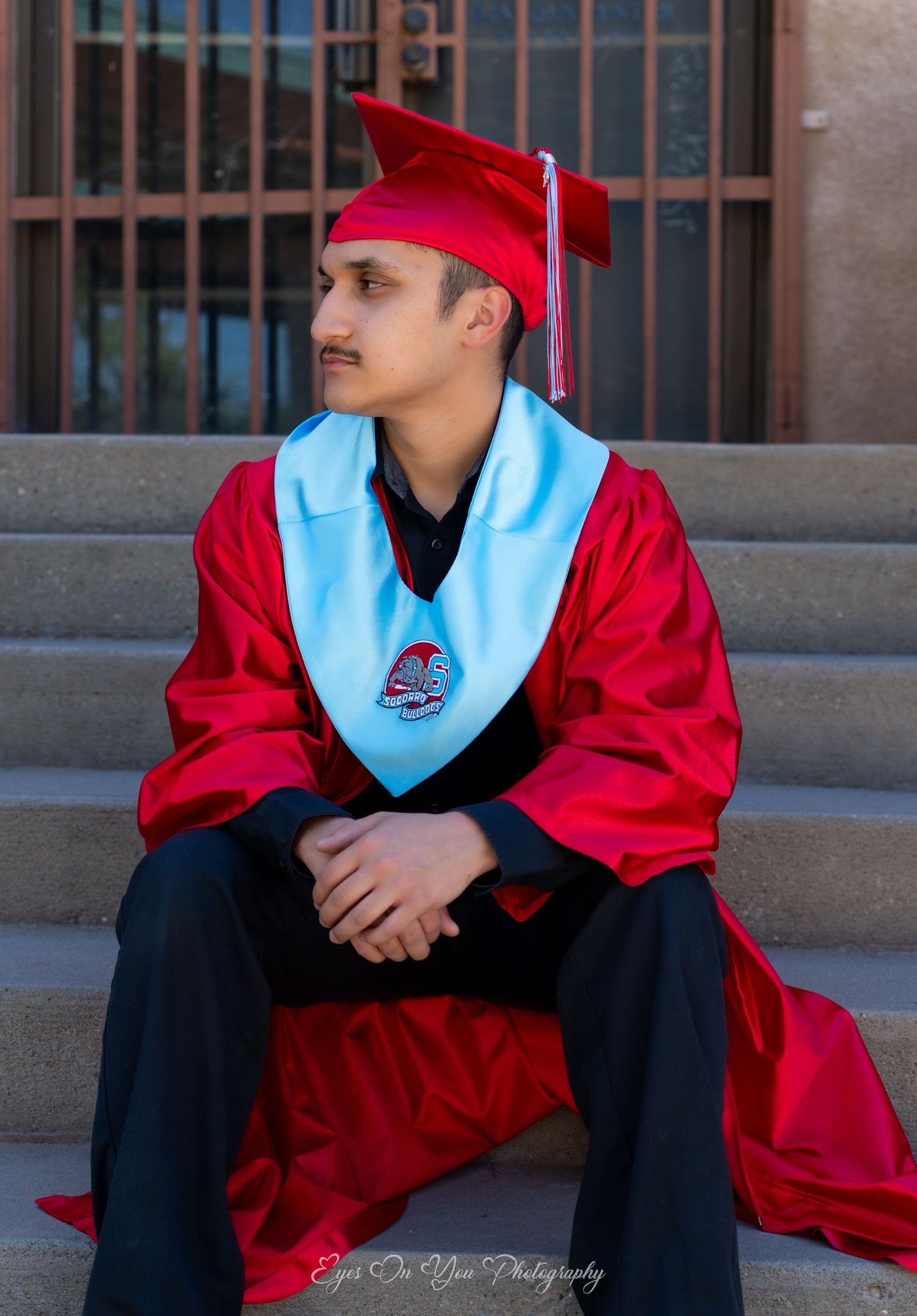 Graduate in red cap and gown with light blue stole sitting thoughtfully.