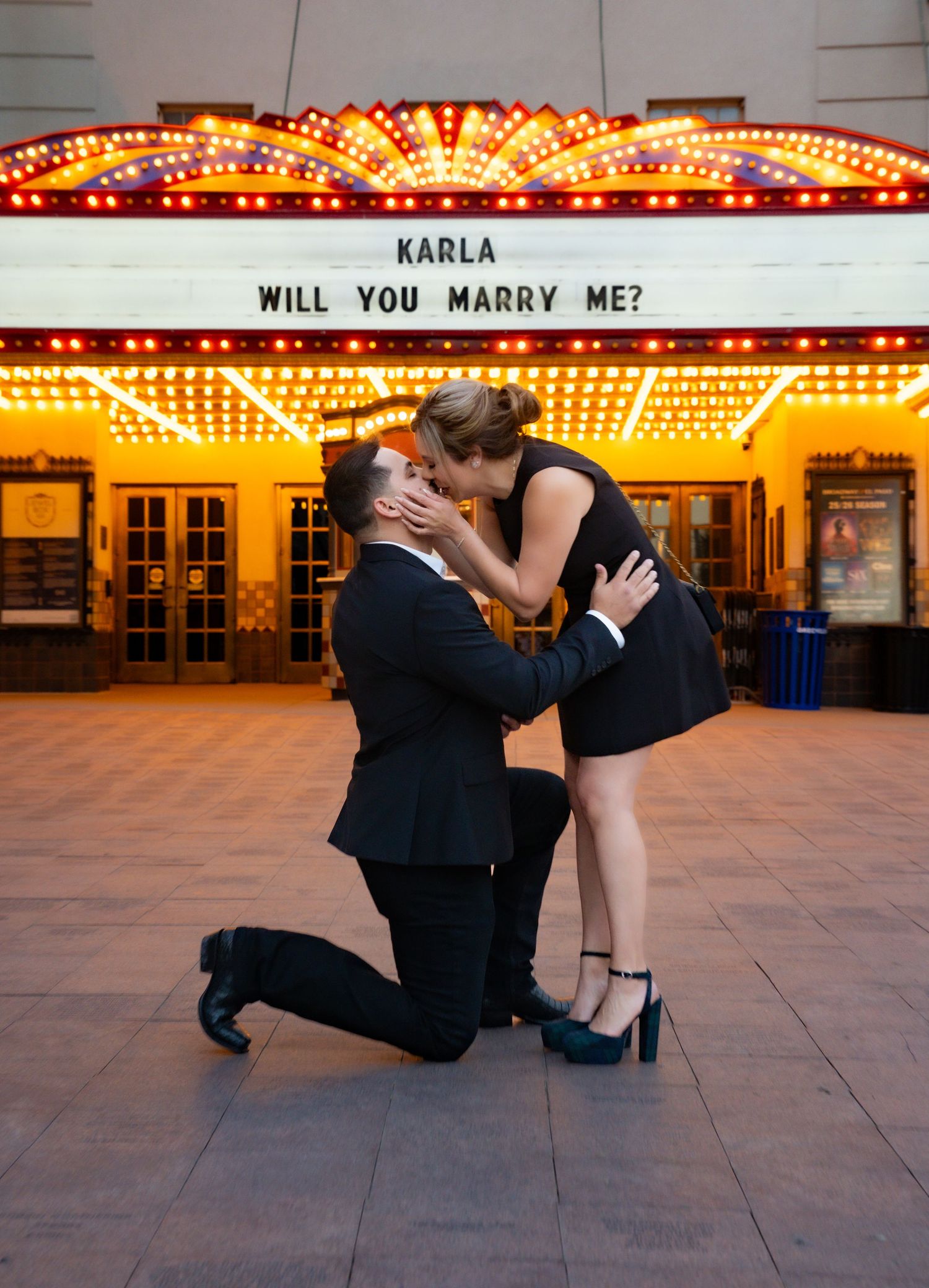 A proposal scene unfolds under a vintage theater marquee with glowing lights at night.