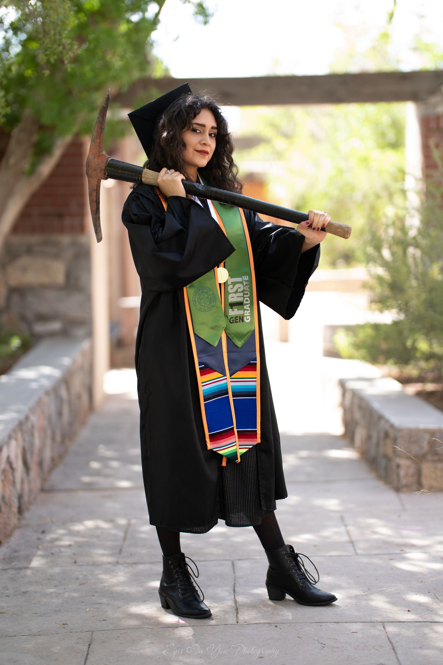 Graduate in black cap and gown displaying a colorful graduation stole against a sunlit outdoor walkway.