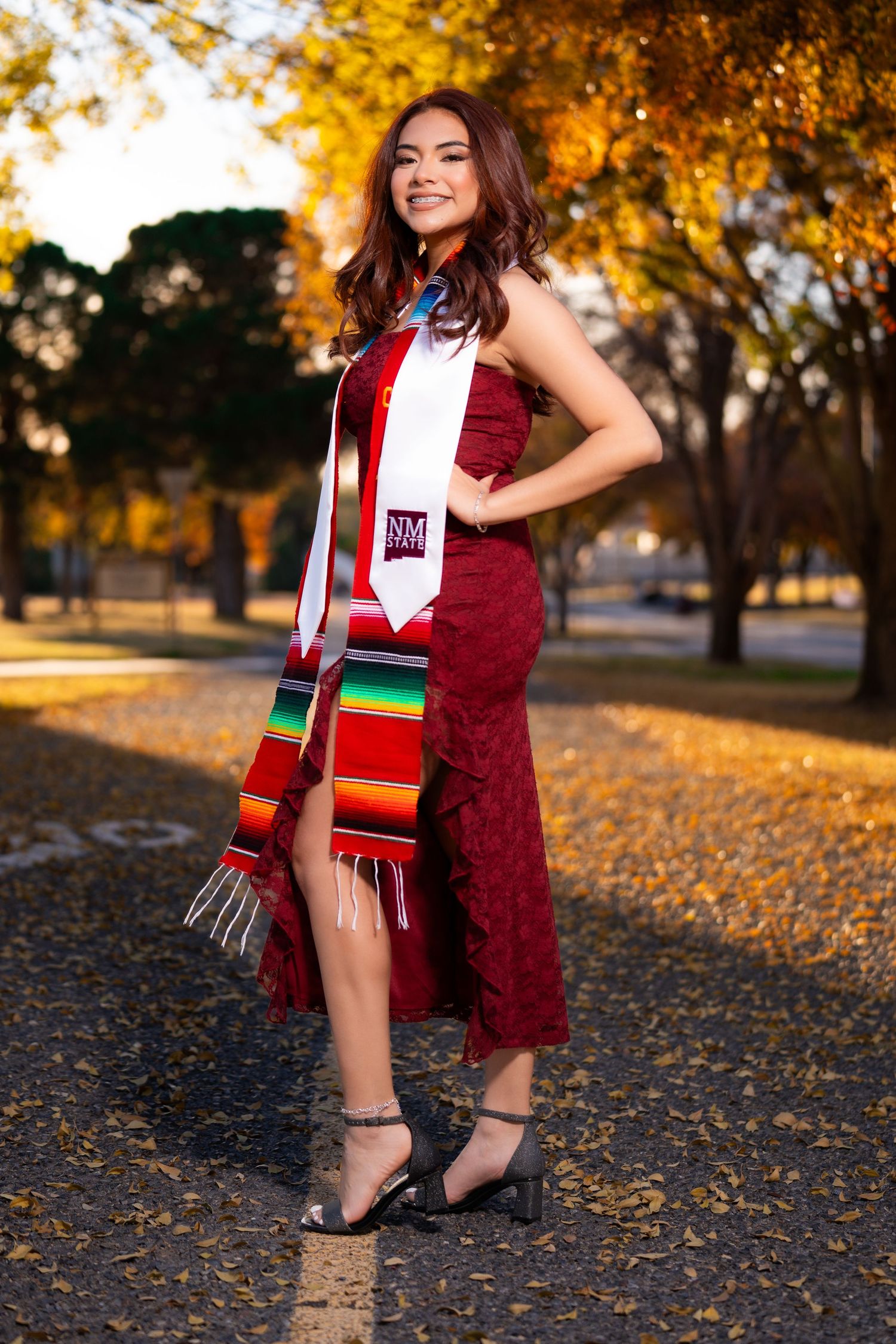 Young woman in maroon graduation dress with NM stole and colorful serape sash posing outdoors in autumn.