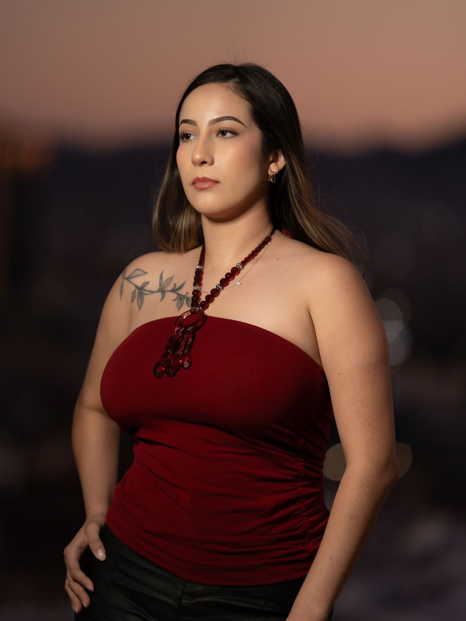 A woman in a red strapless top poses outdoors at sunset with a beaded necklace and visible shoulder tattoo.