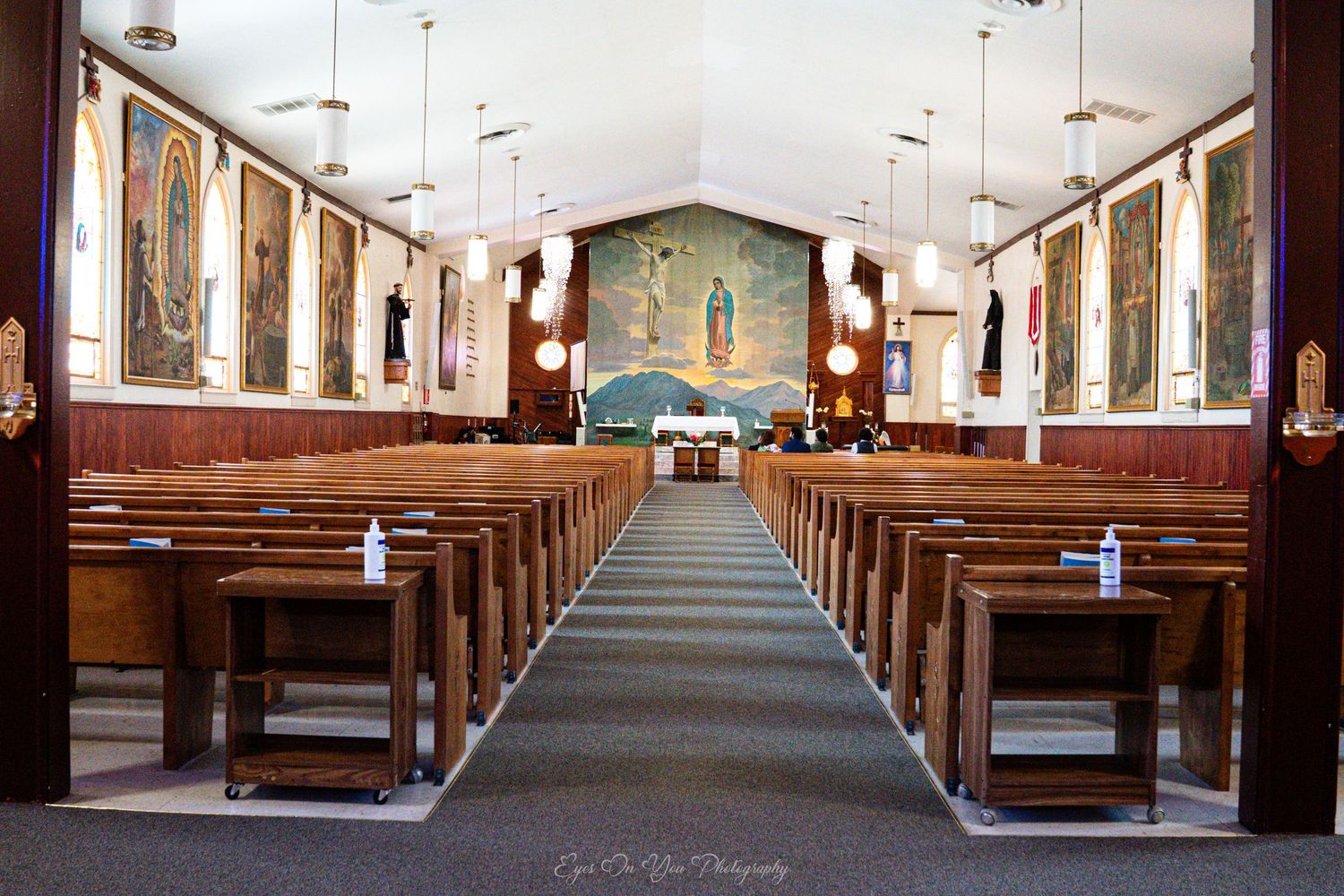 Interior view of empty wooden church pews in a Catholic church sanctuary with religious artwork on walls.
