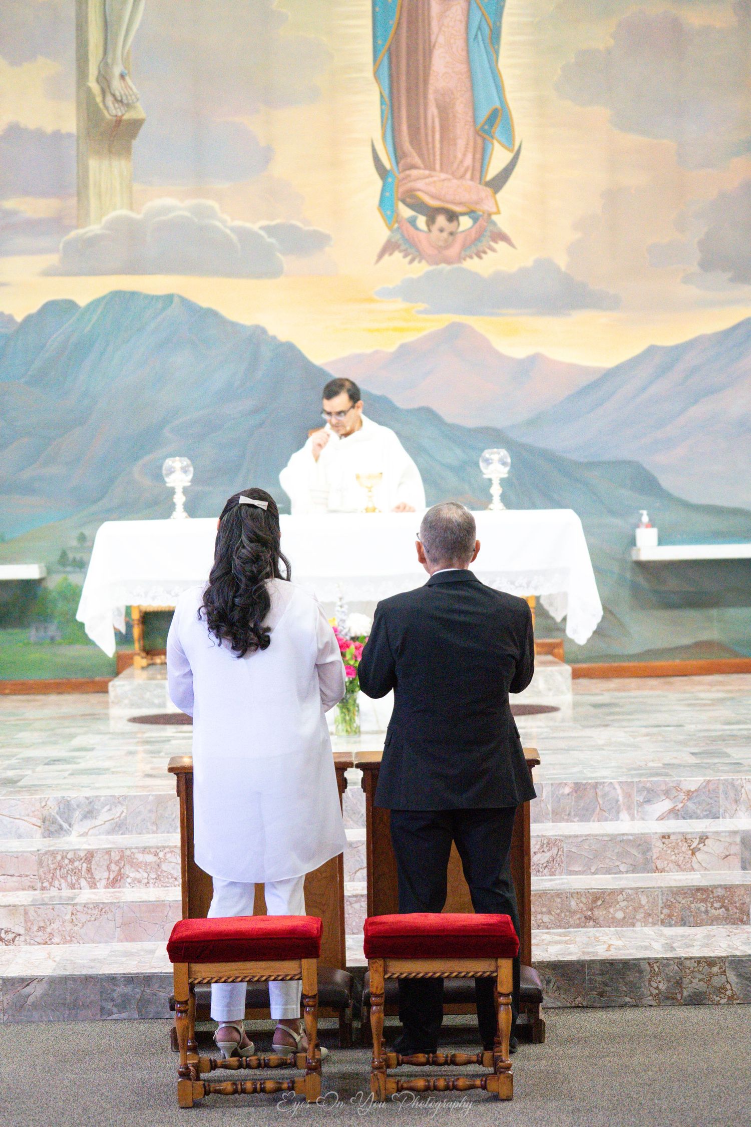 Bride and groom kneel at church altar during Catholic wedding ceremony with mountain landscape mural in background.