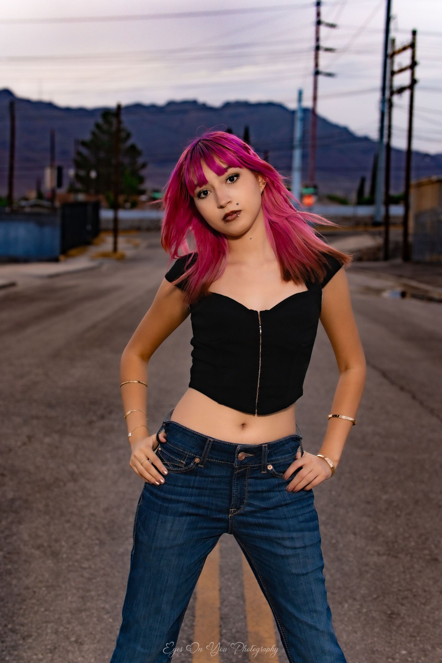 Standing on a road with mountains behind, wearing black crop top and jeans with vibrant pink hair.