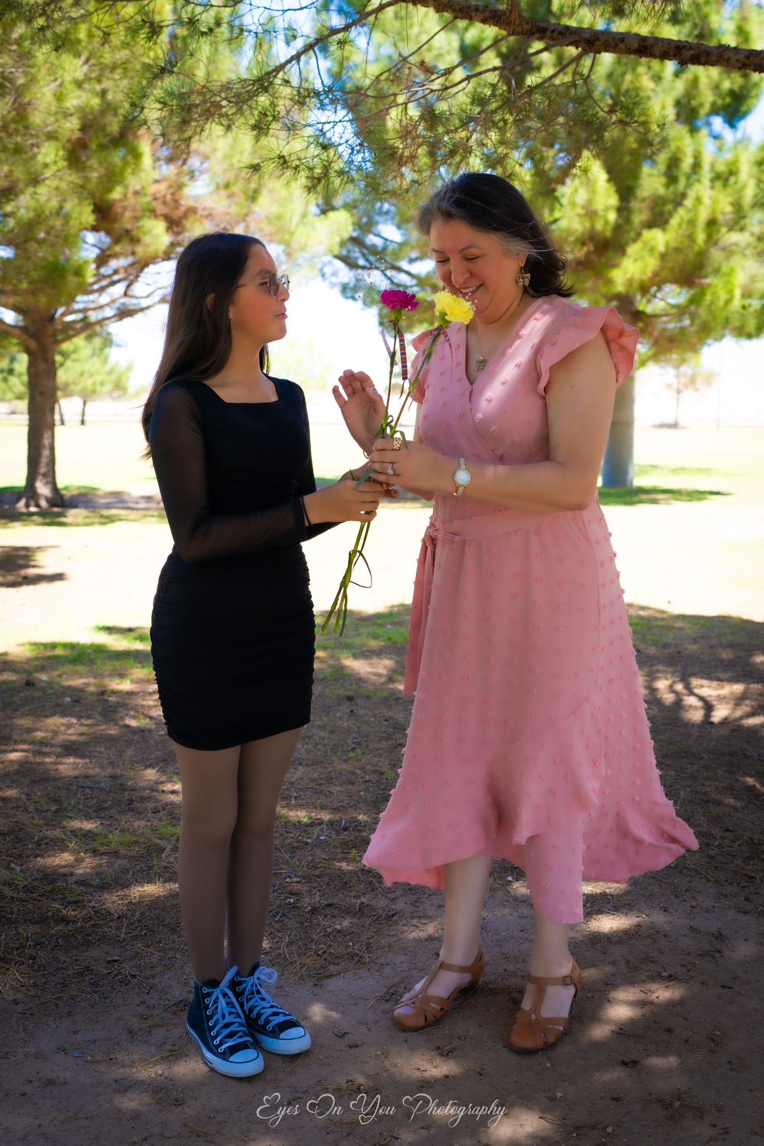 Two figures in contrasting black and pink outfits stand together under pine trees in a park.