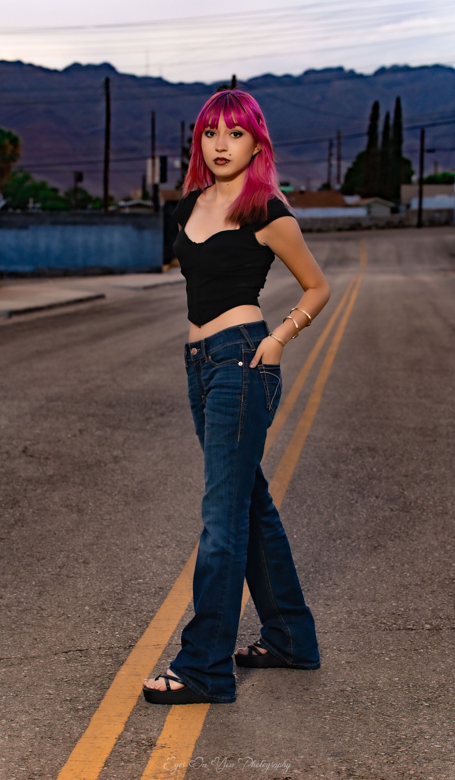 Sunset street fashion photo with pink hair, black top and flared jeans against mountain backdrop.