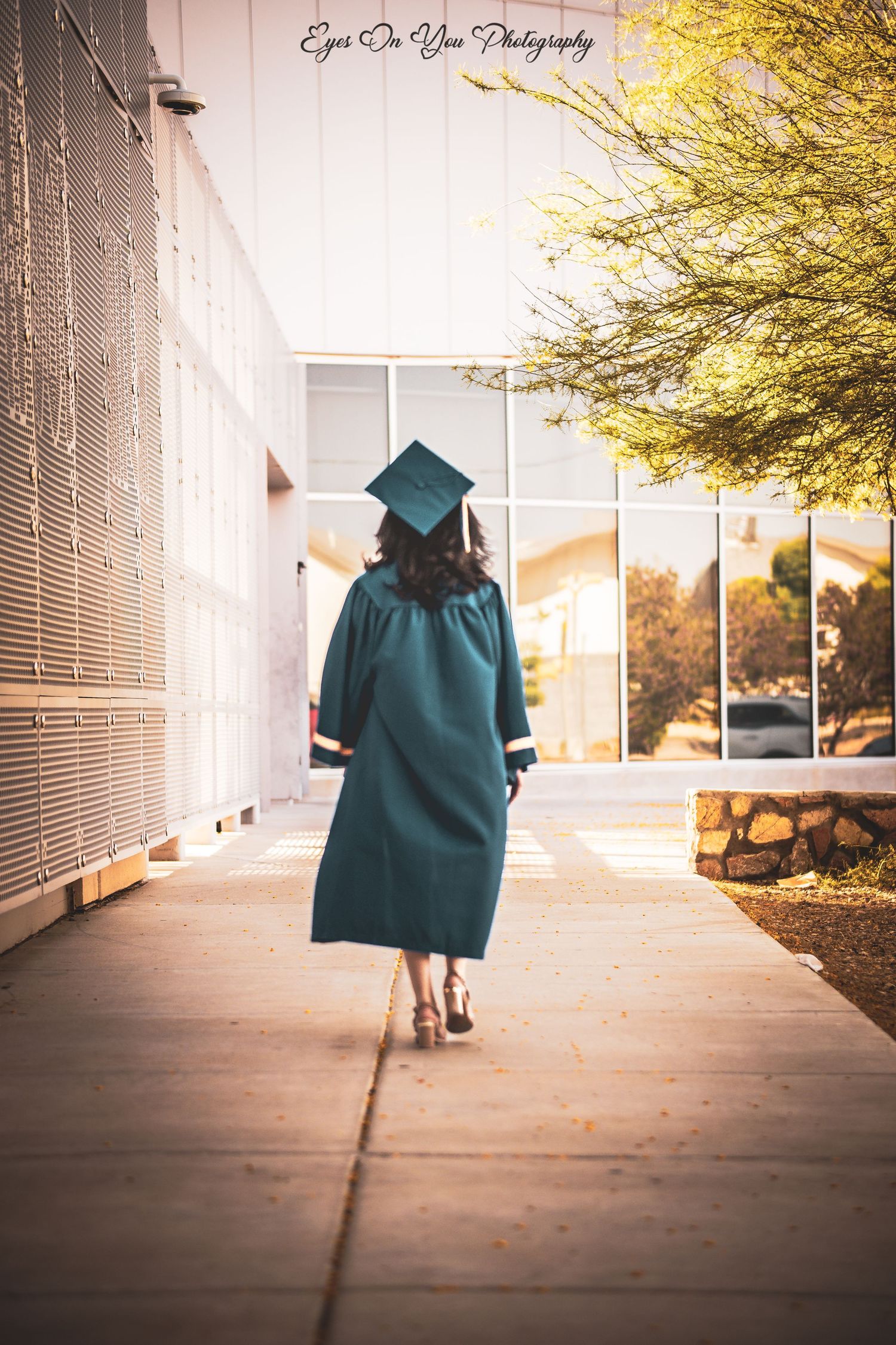 Graduate in a teal cap and gown walking away on a concrete path during golden hour.