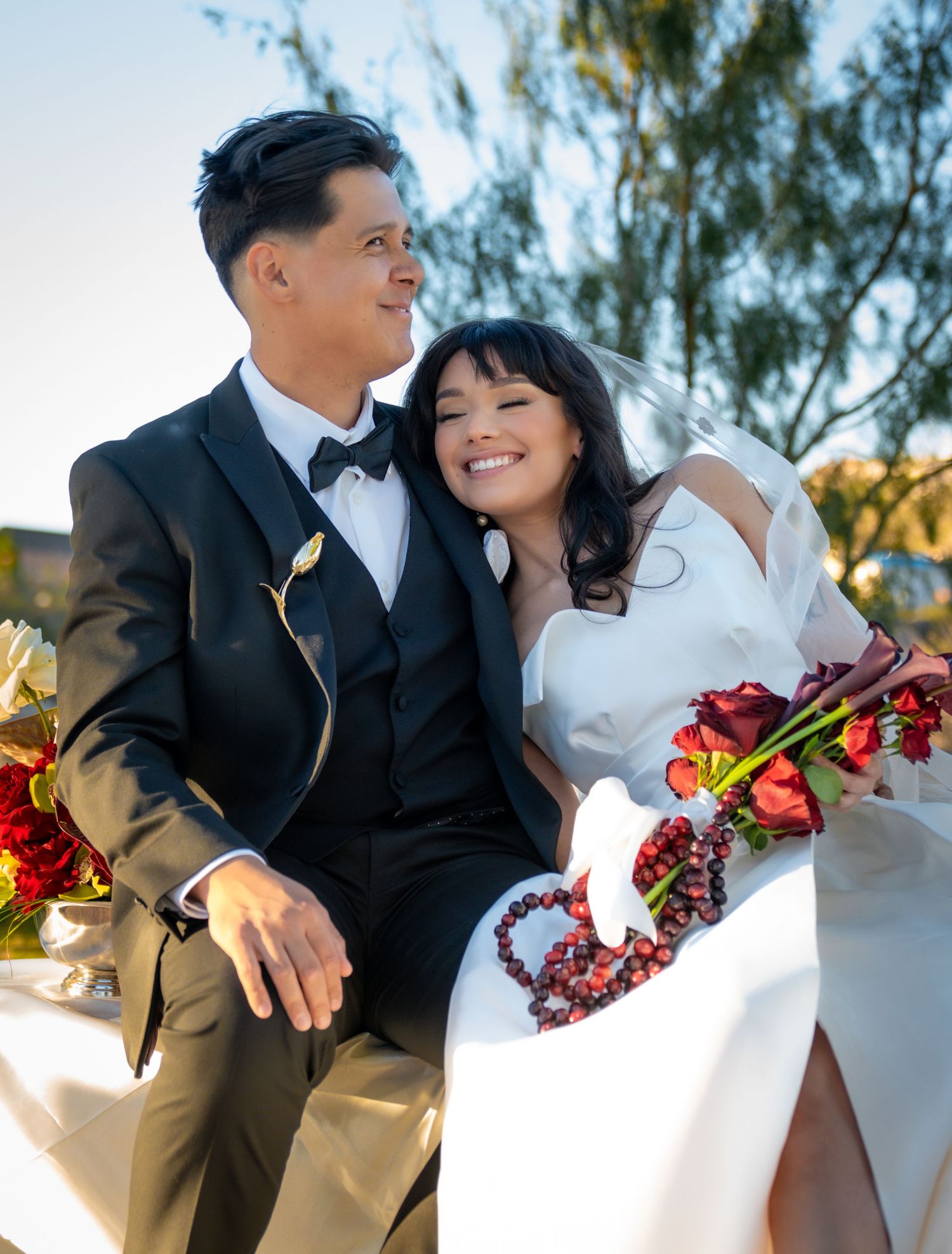 A couple shares a romantic moment in formal wedding attire with red floral bouquet against a sunny outdoor backdrop.