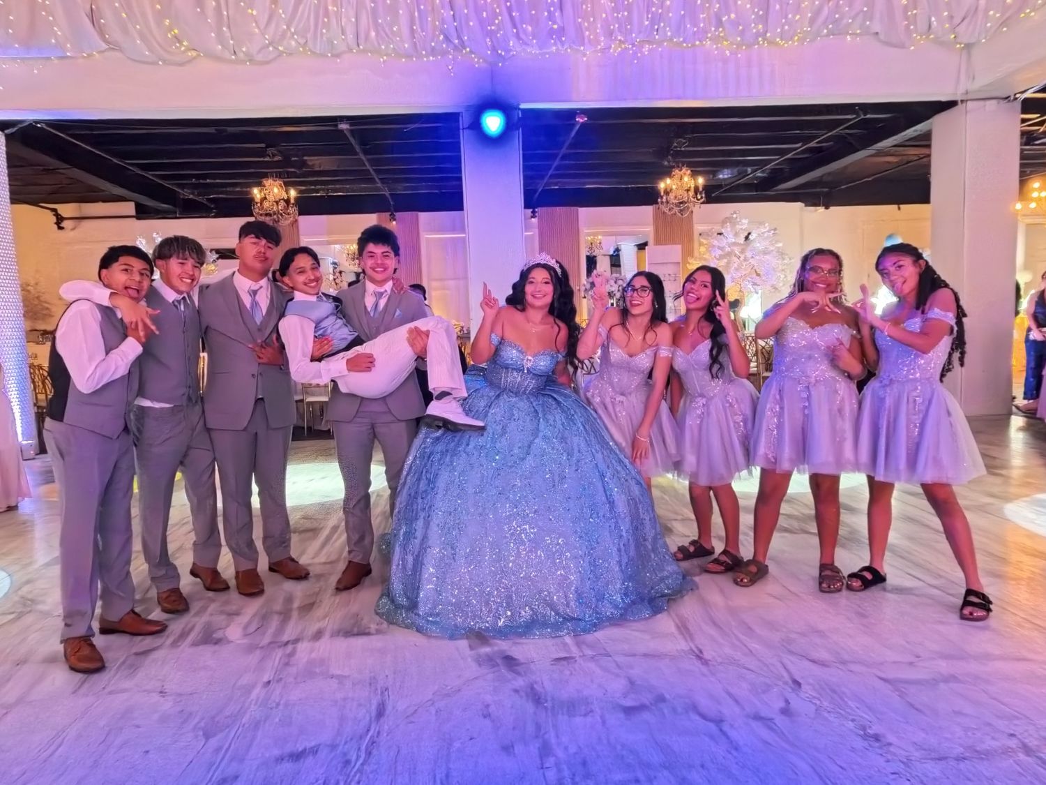 Teen girl in blue quinceañera gown poses with court of honor in gray suits and lilac dresses at elegant venue.
