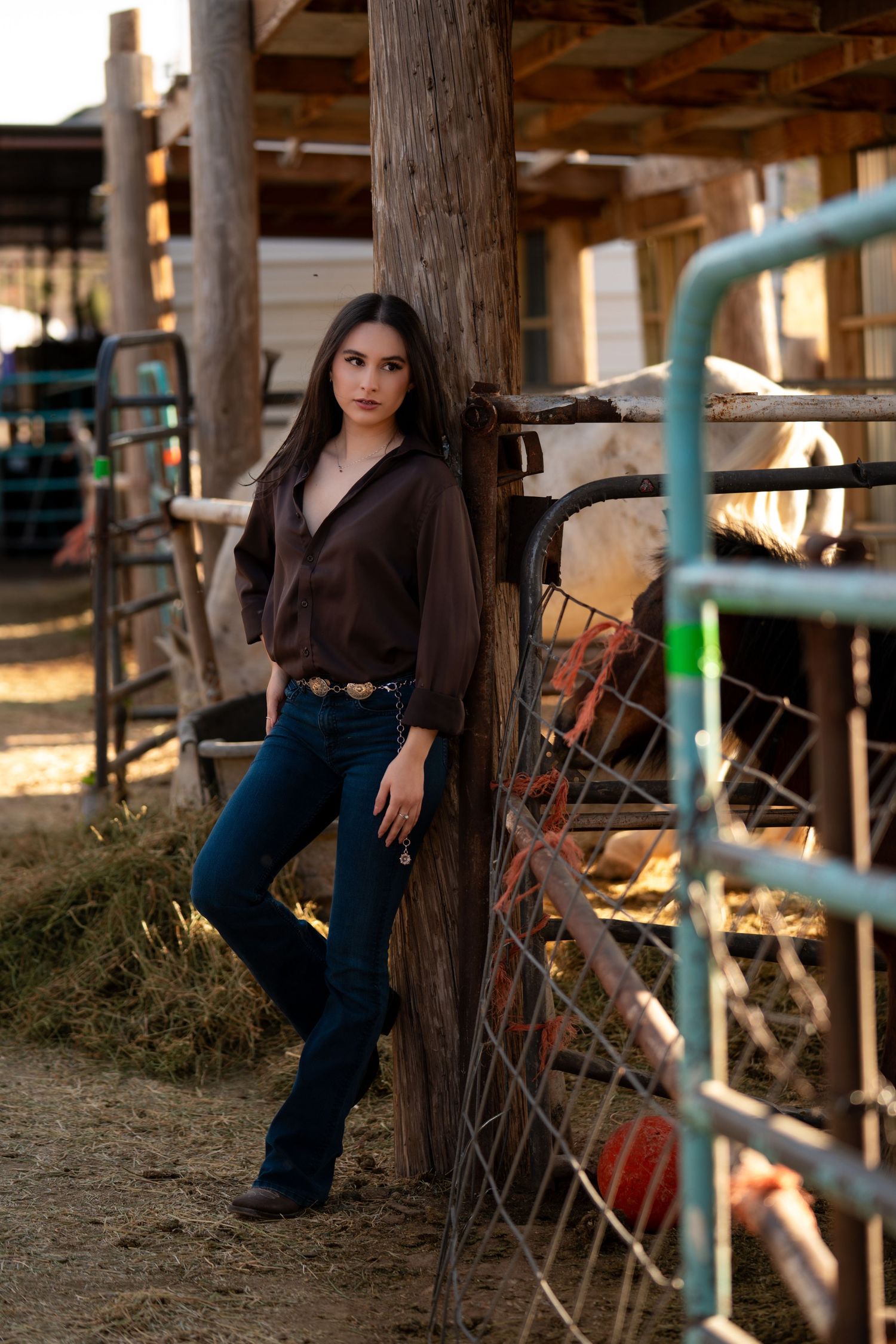 A person in a dark top and jeans poses near rustic wooden beams and metal railings in a barn-like setting at sunset.