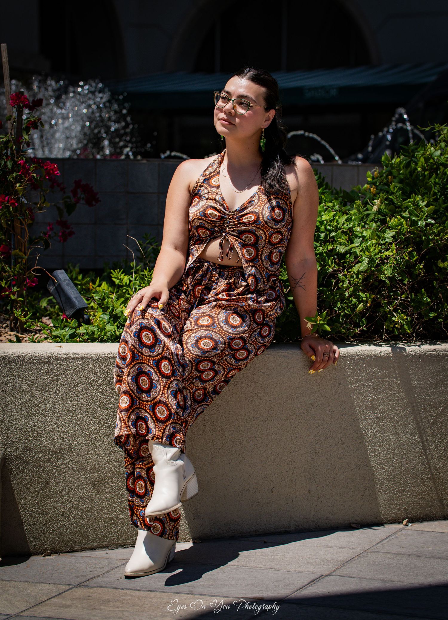 Person in patterned dress sits casually on concrete ledge with greenery in background.