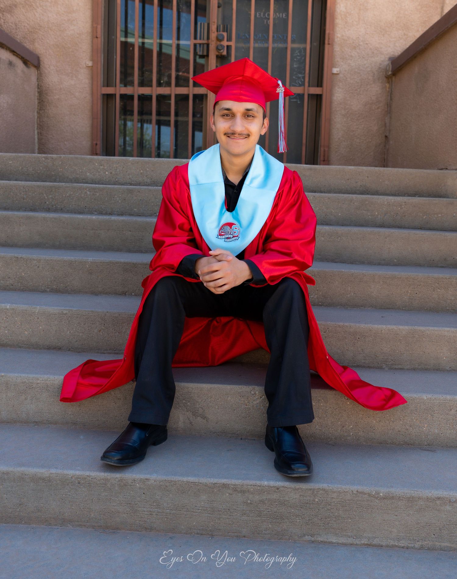 Graduate in red cap and gown sits casually on concrete steps during graduation photoshoot.