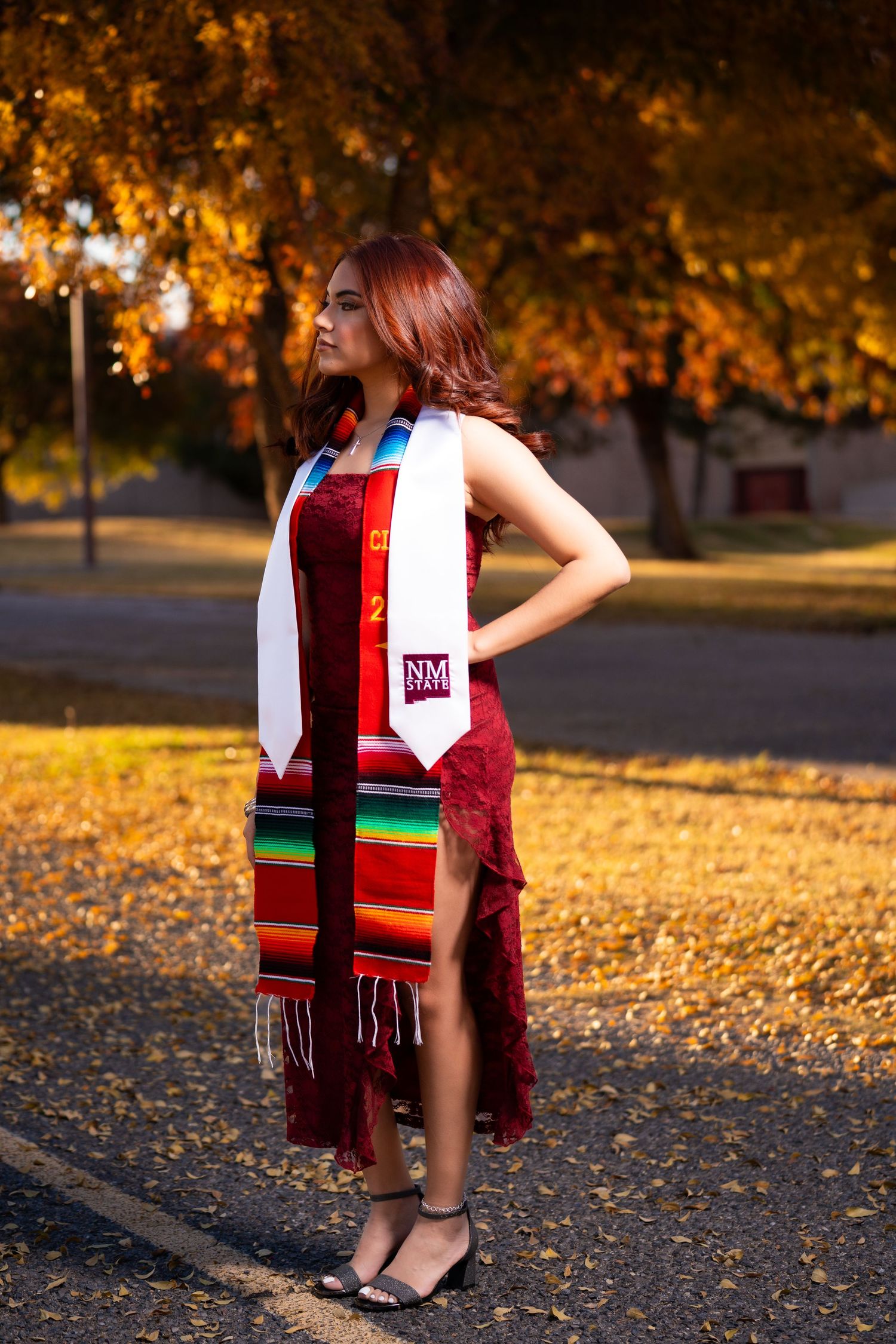 A graduate in a red dress and colorful graduation stole poses outdoors during golden hour with autumn leaves in background.