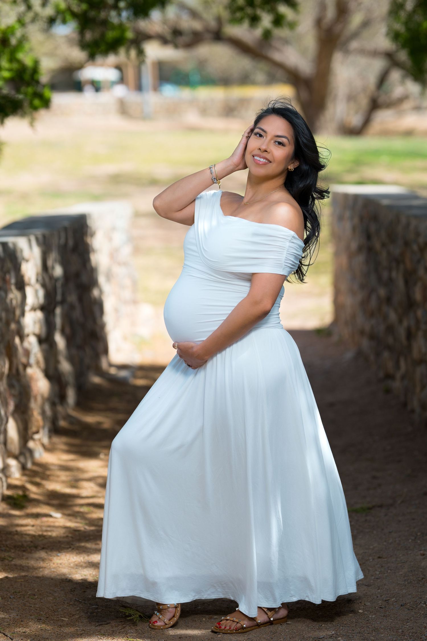 Smiling pregnant woman in white off-shoulder maxi dress cradling baby bump outdoors in a park setting.