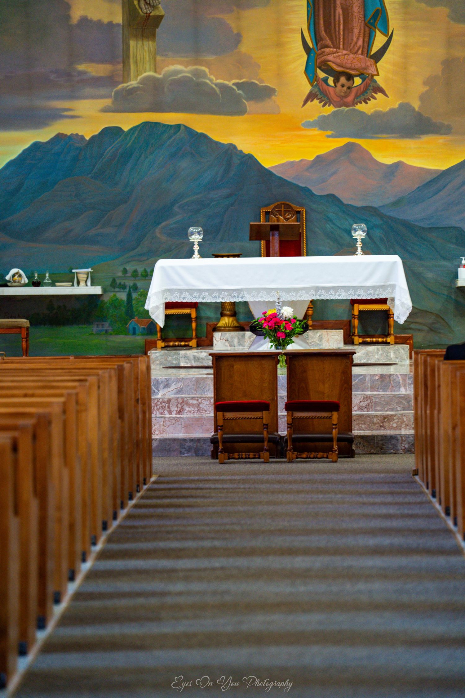 Inside a rustic church with wooden pews leading to an altar decorated with white linens against a painted mountain backdrop.