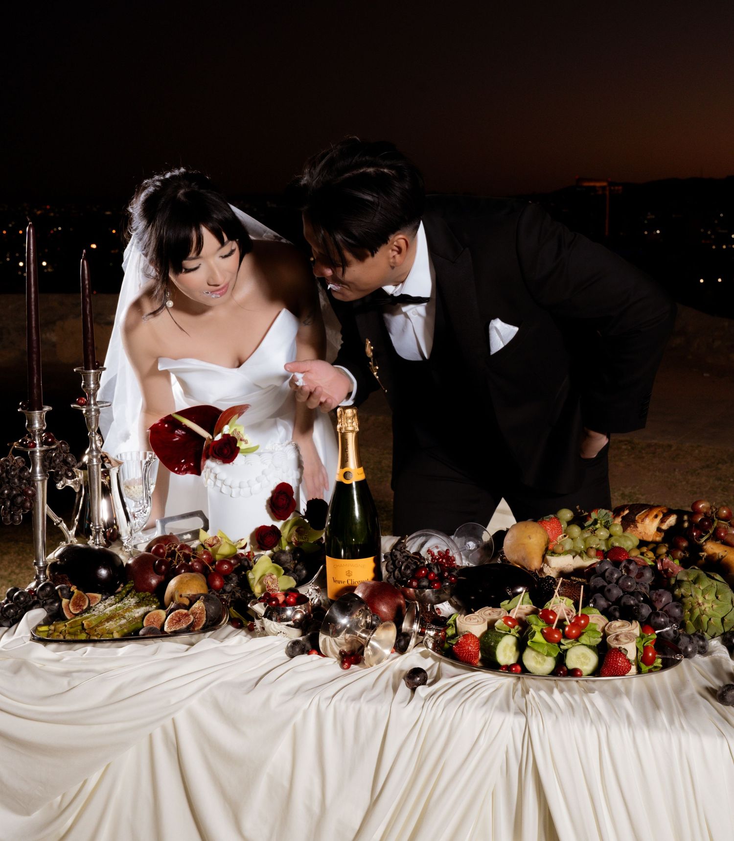 A wedding reception table features an elegant spread of fruits, flowers, and wine bottles in a dark, moody setting.