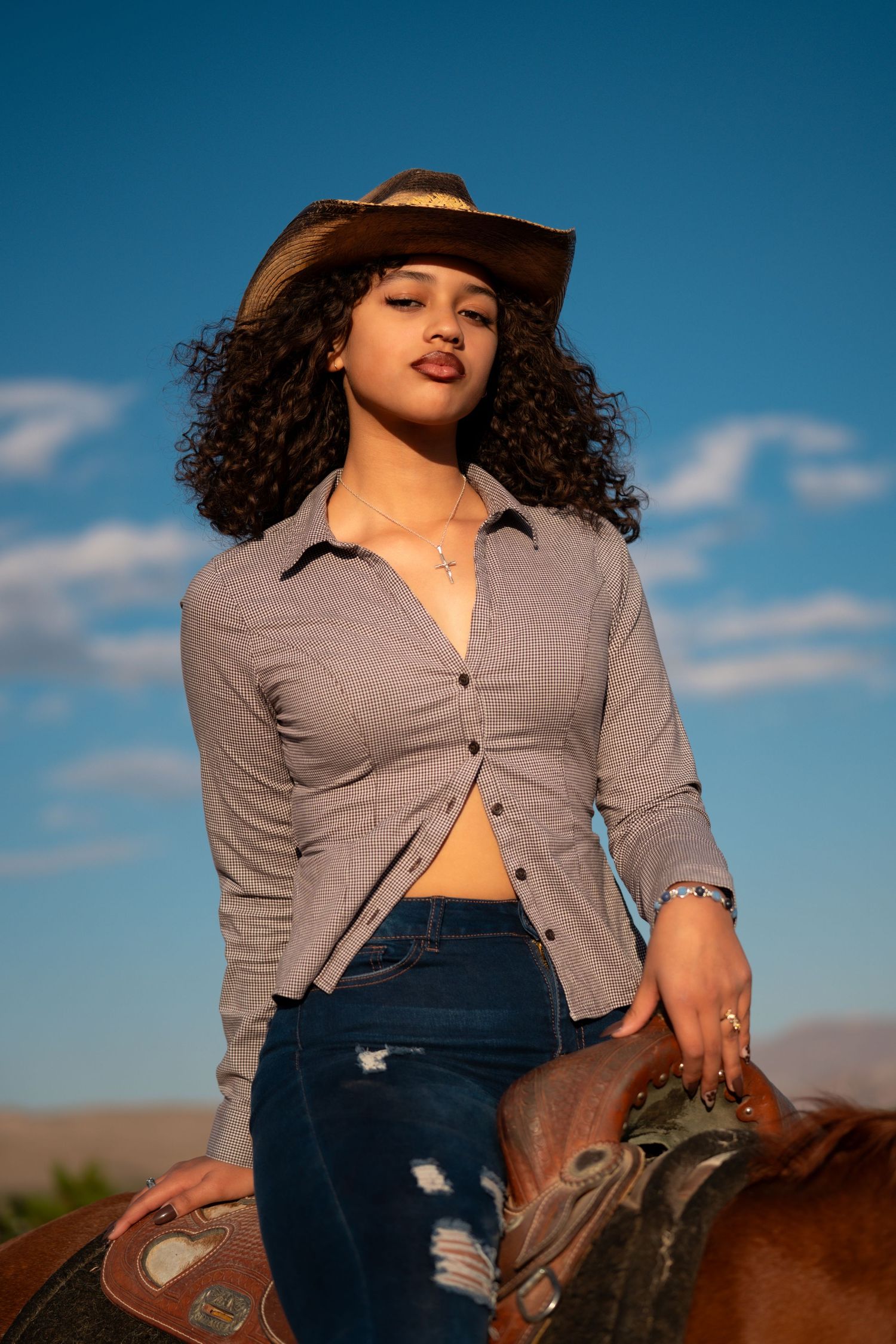 A woman in a gray button-up shirt and jeans rides horseback against a bright blue sky during golden hour.
