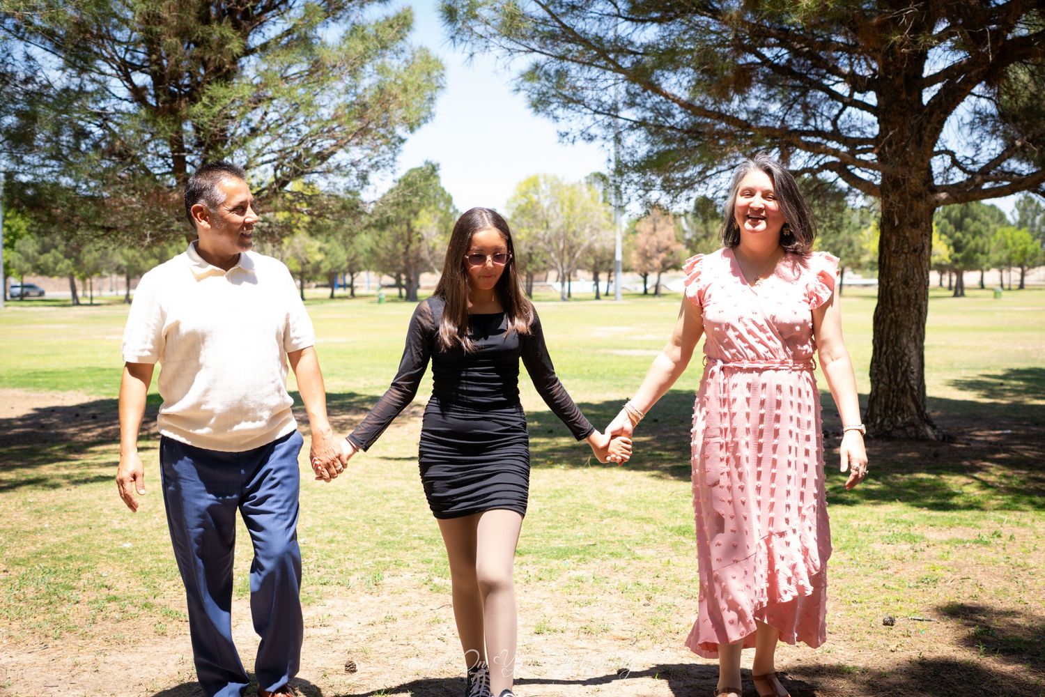 Three people hold hands while walking together in a sunny park with pine trees.