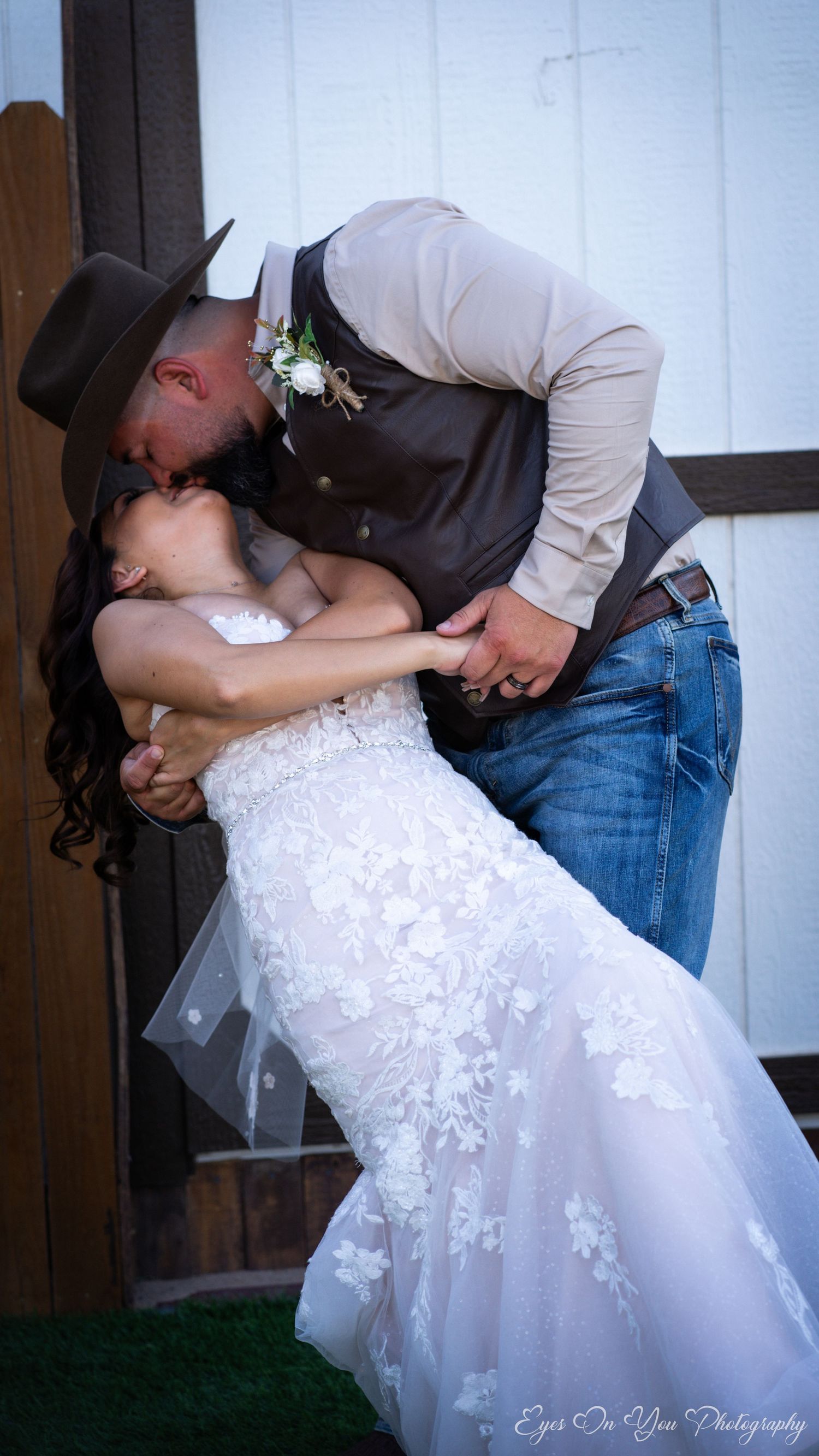 Romantic couple shares a dip kiss while dressed in wedding attire against a rustic wall backdrop.